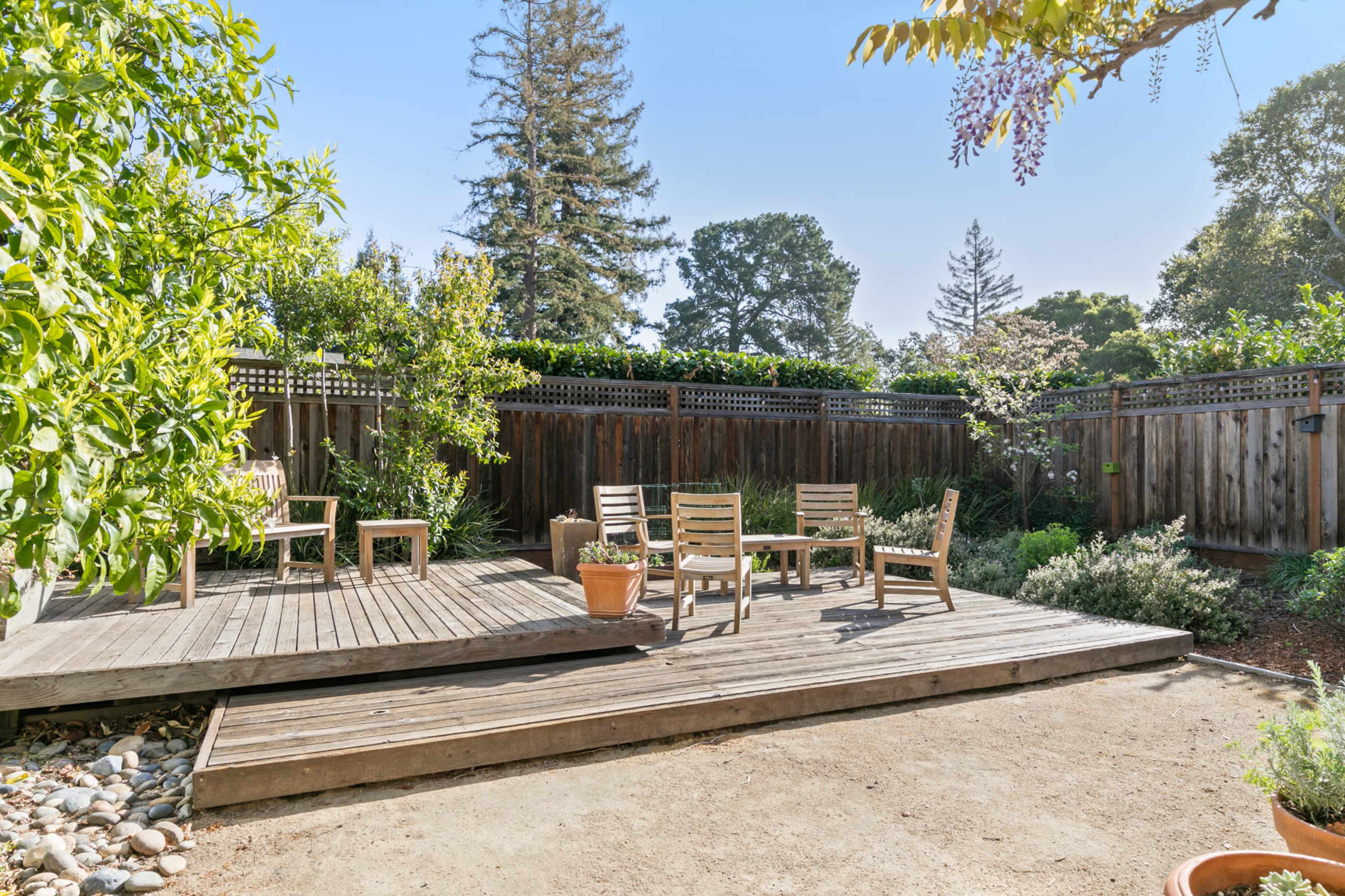A wooden deck in a garden surrounded by trees and plants, with chairs and a table arranged on the deck.
