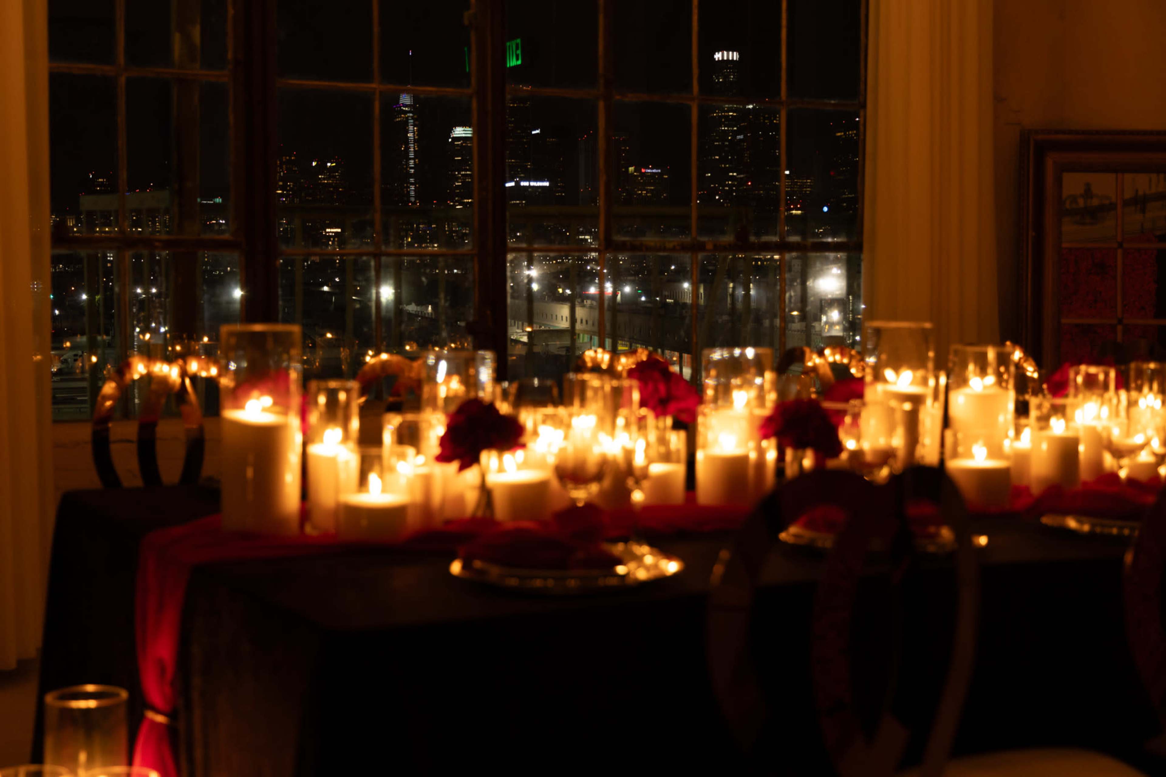 A dimly lit dining table adorned with candles and red flowers, overlooking a city skyline at night through large windows.