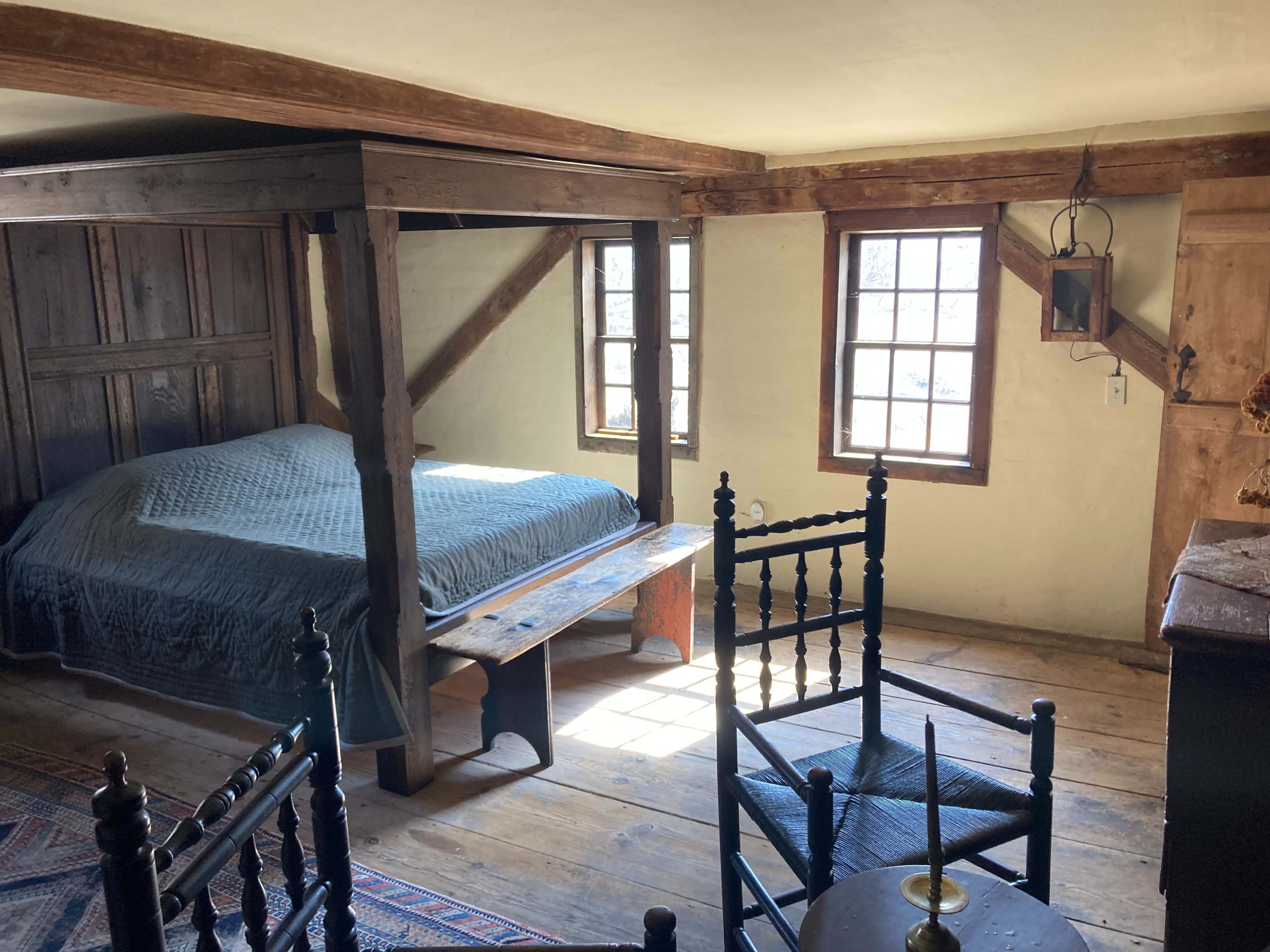 A rustic bedroom with a wooden bed frame, a table, chairs, and windows allowing natural light to enter.