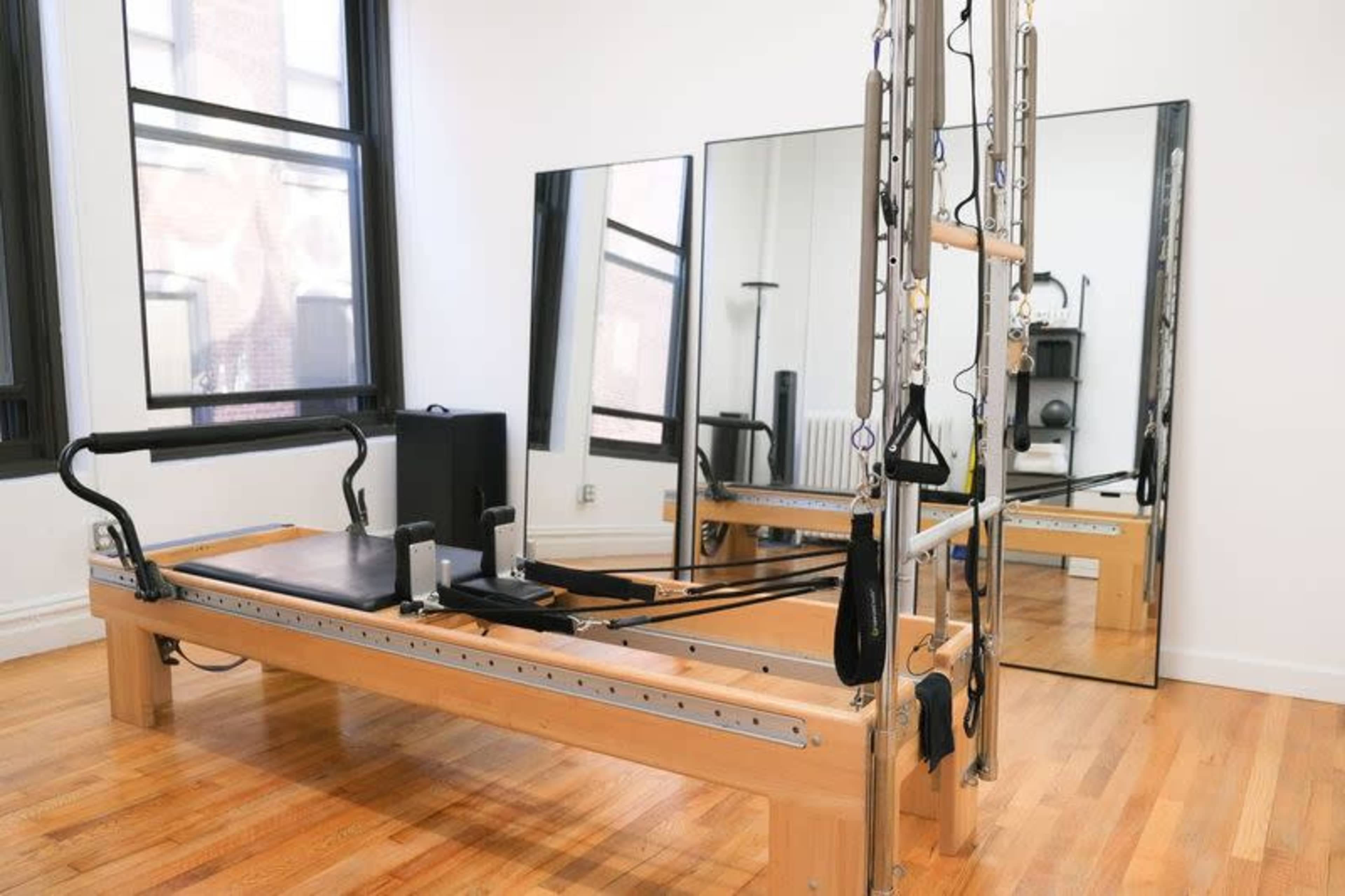 The image shows a Pilates reformer positioned in a bright, airy room with large mirrors and hardwood flooring.
