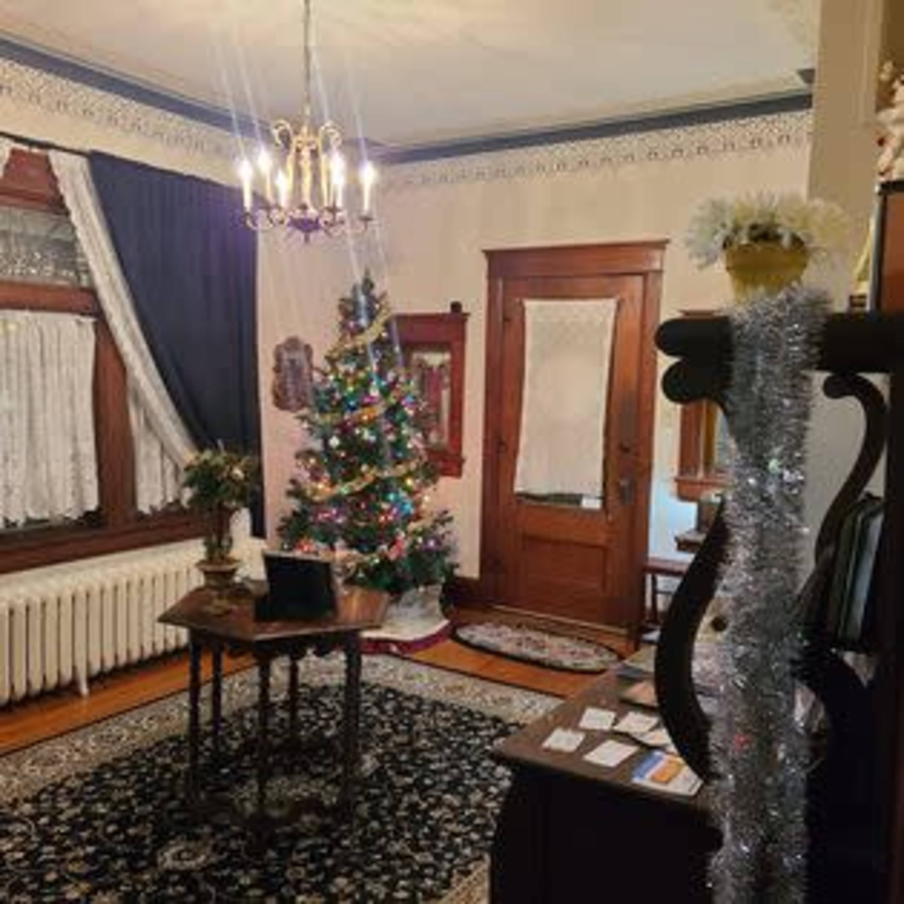 The room features a decorated Christmas tree, vintage furnishings, and an ornate chandelier, with a large area rug on the floor.