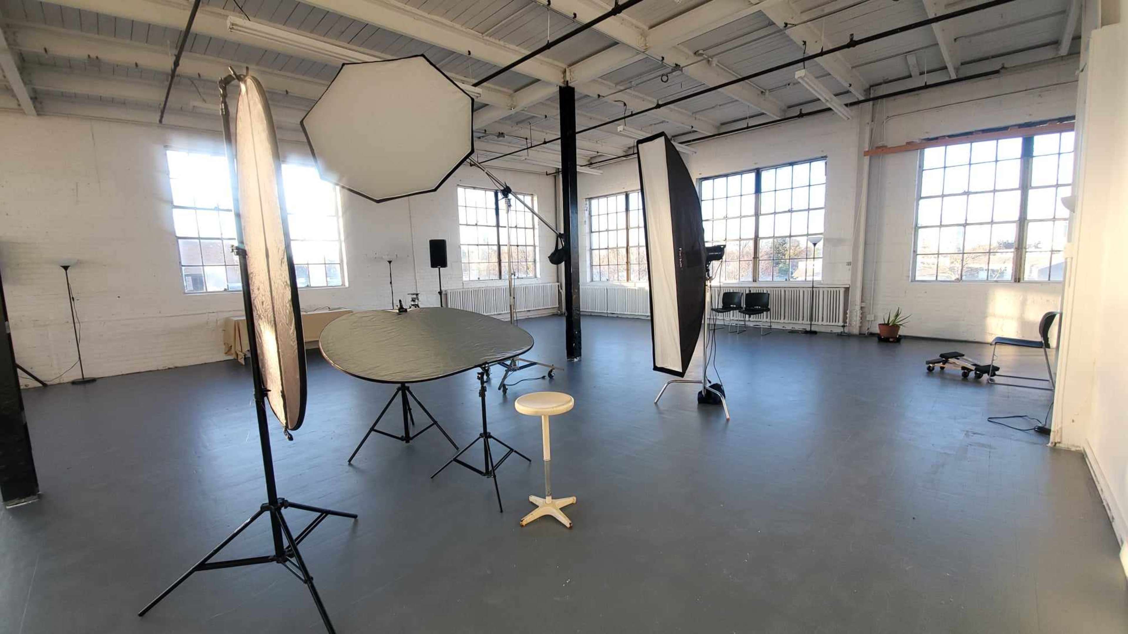 An empty photography studio with various lighting equipment and a stool arranged in a spacious room filled with natural light.