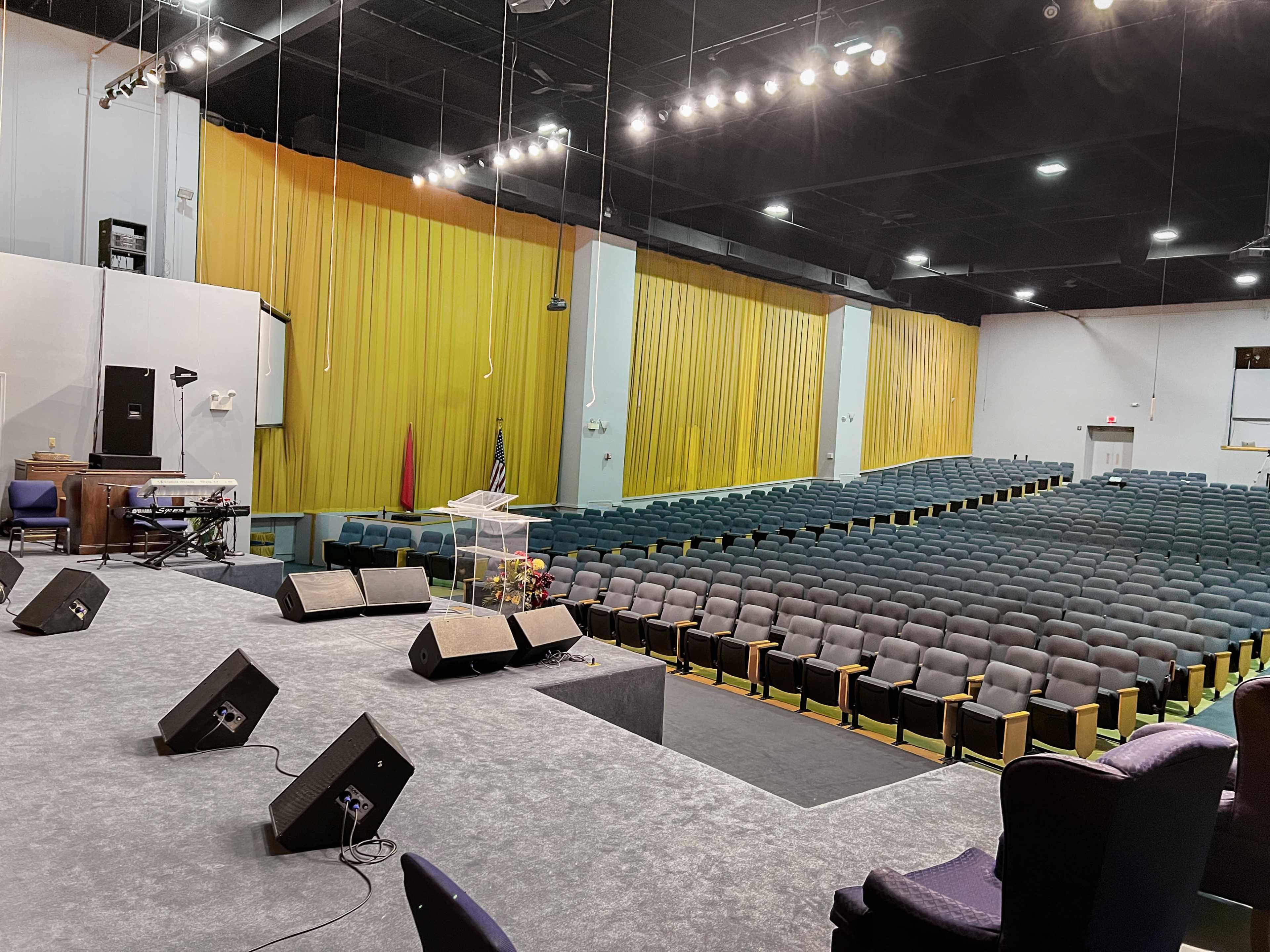 An empty auditorium with rows of seats and a stage featuring a clear podium and microphones.