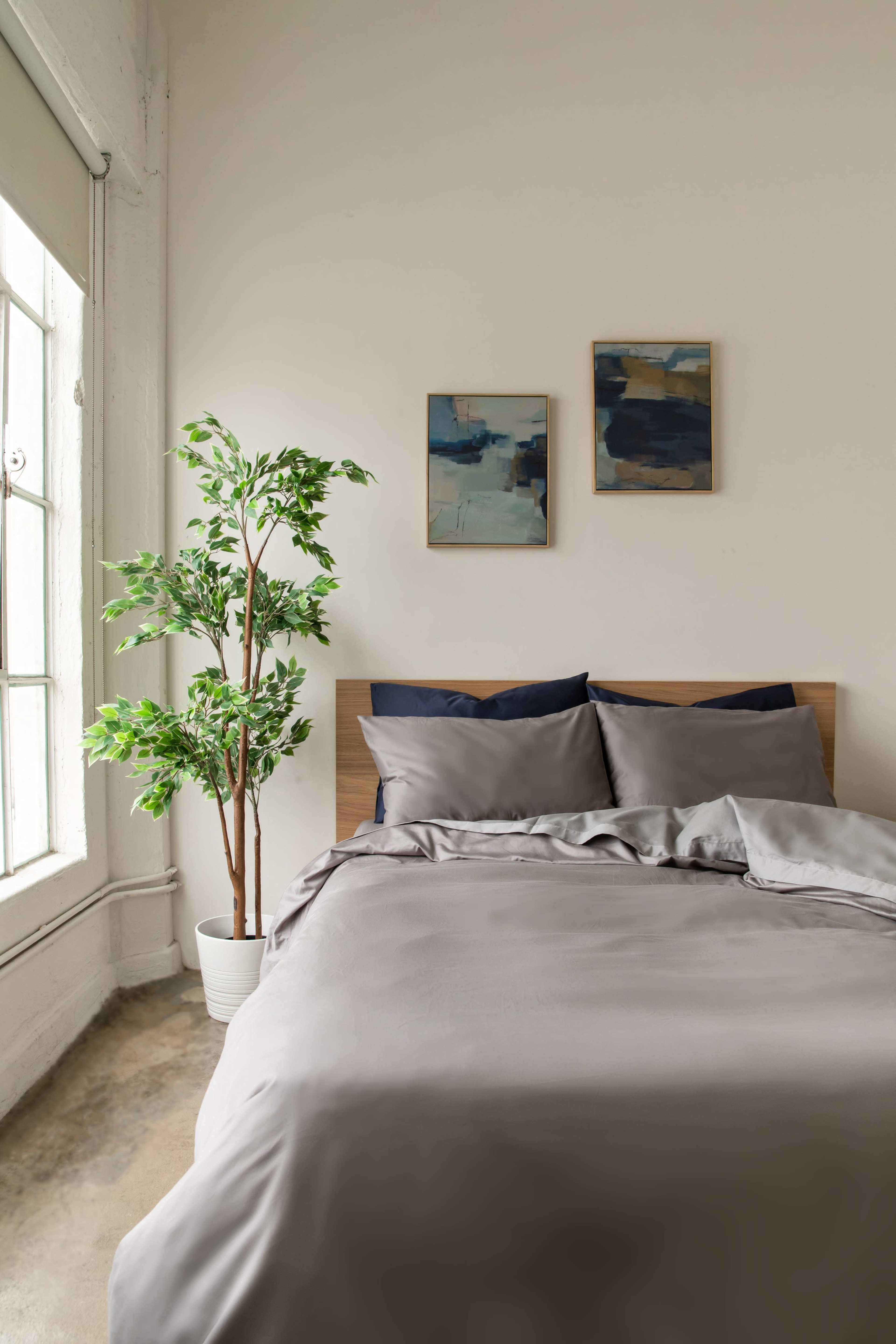 A neatly made bed with gray linens is positioned against a wall, accompanied by two framed artworks and a potted plant near a large window.