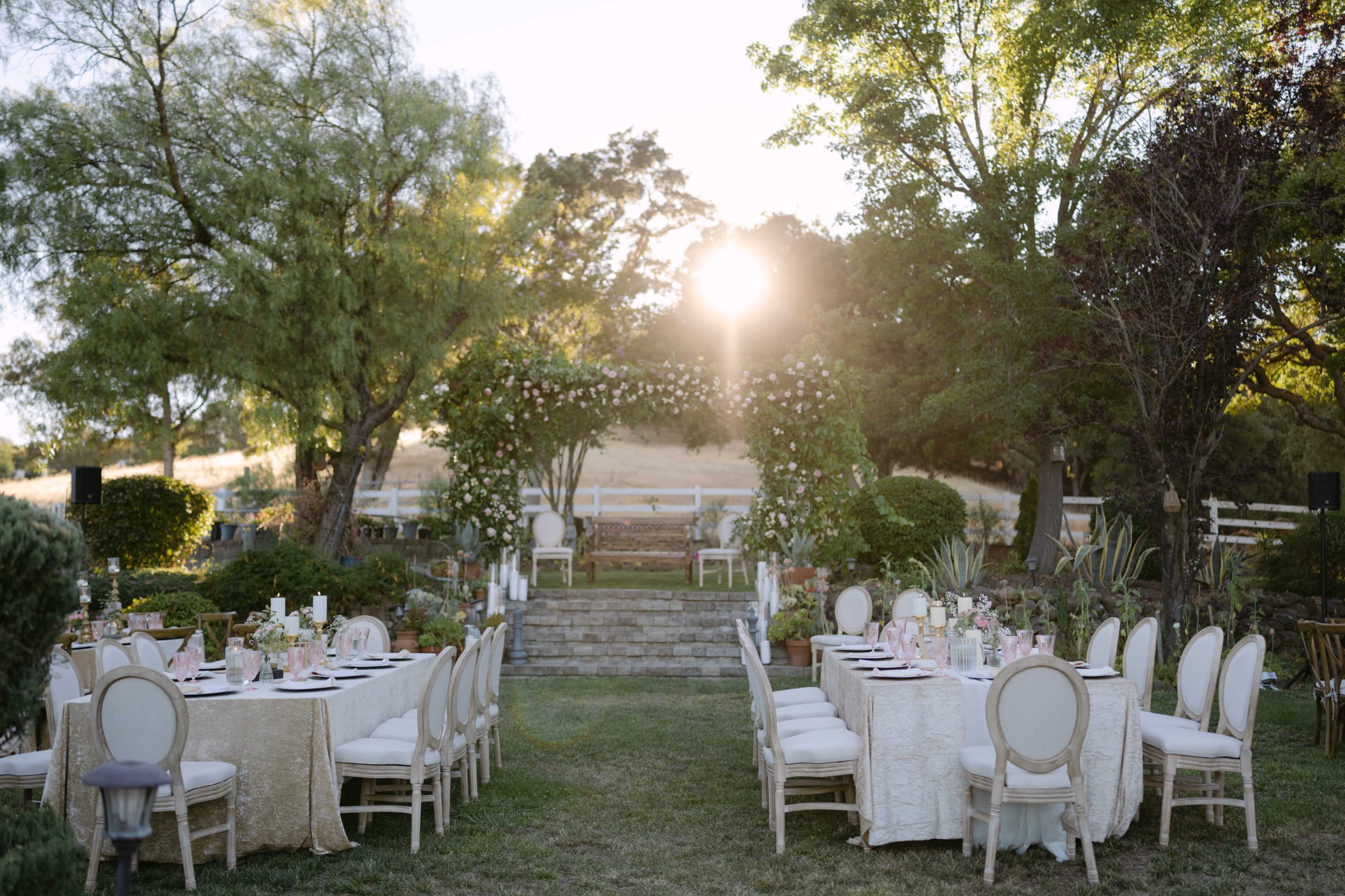 A beautifully arranged outdoor setting features long tables with elegant decorations, surrounded by greenery and a wooden archway, with the sun setting in the background.