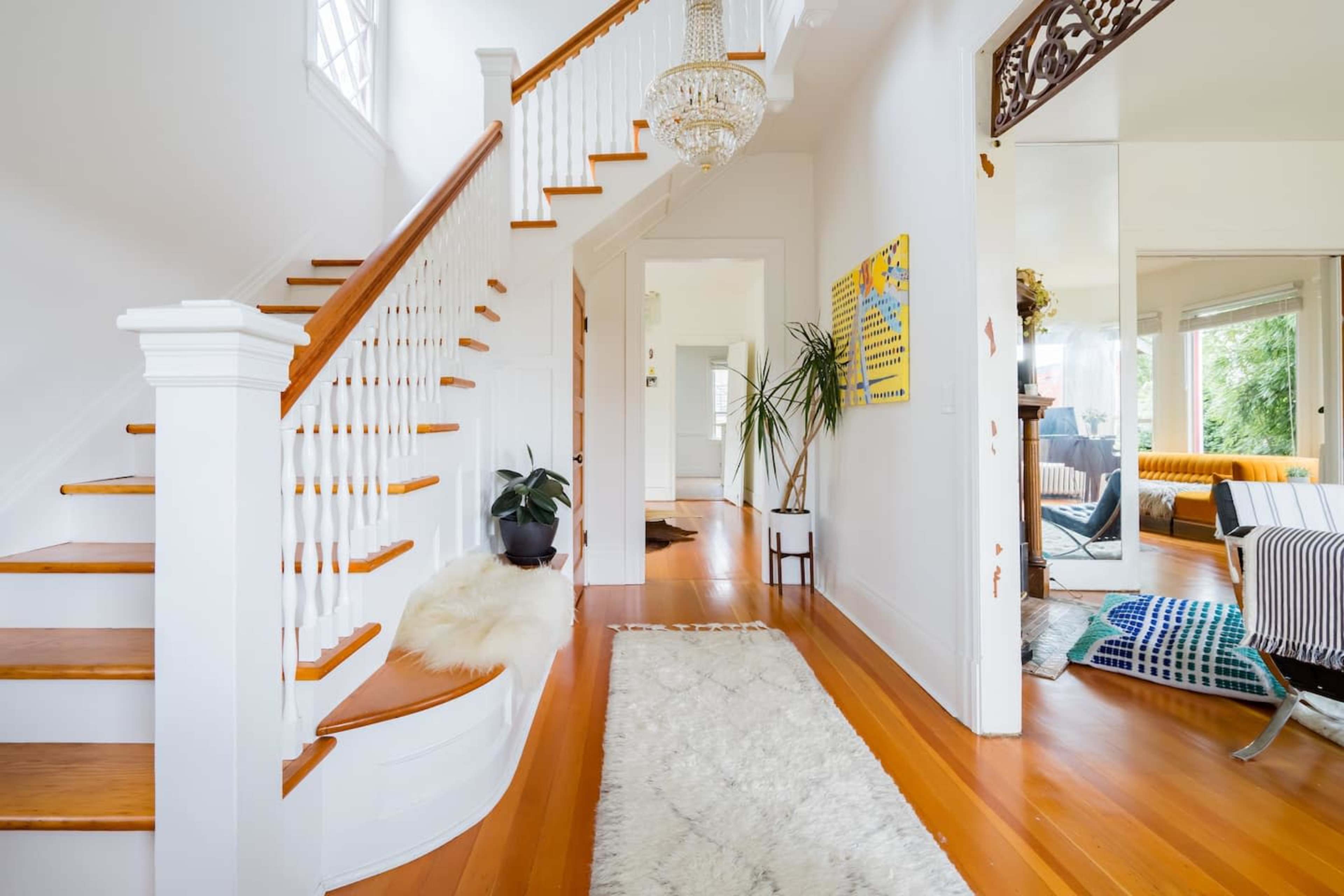 A bright hallway features a wooden staircase, a chandelier, and a large mirror, with a potted plant and a soft rug on the polished floor.