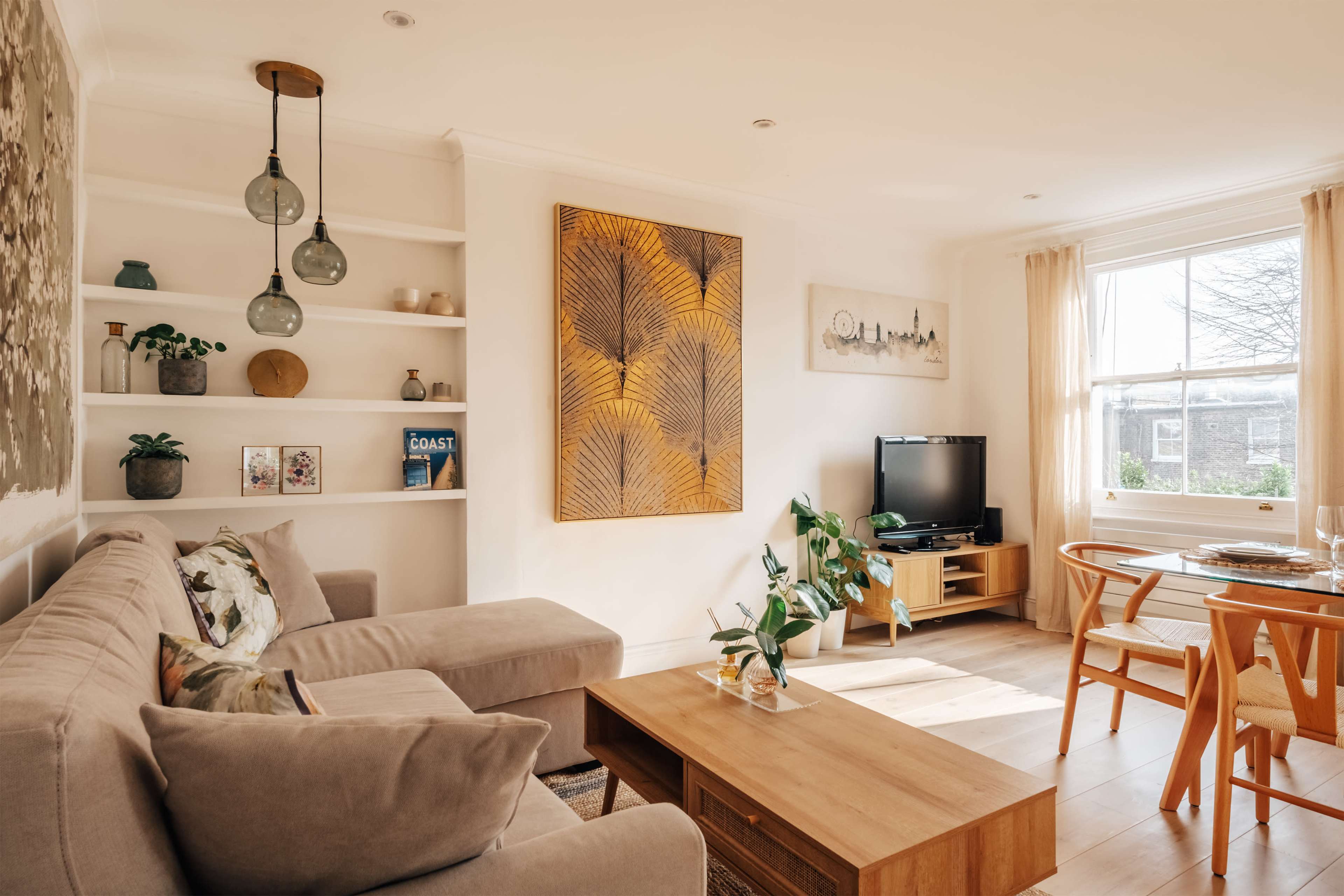 A cozy living room features a light-colored sofa, wooden coffee table, and a television on a media unit, with a large artwork and plants decorating the walls and shelves.