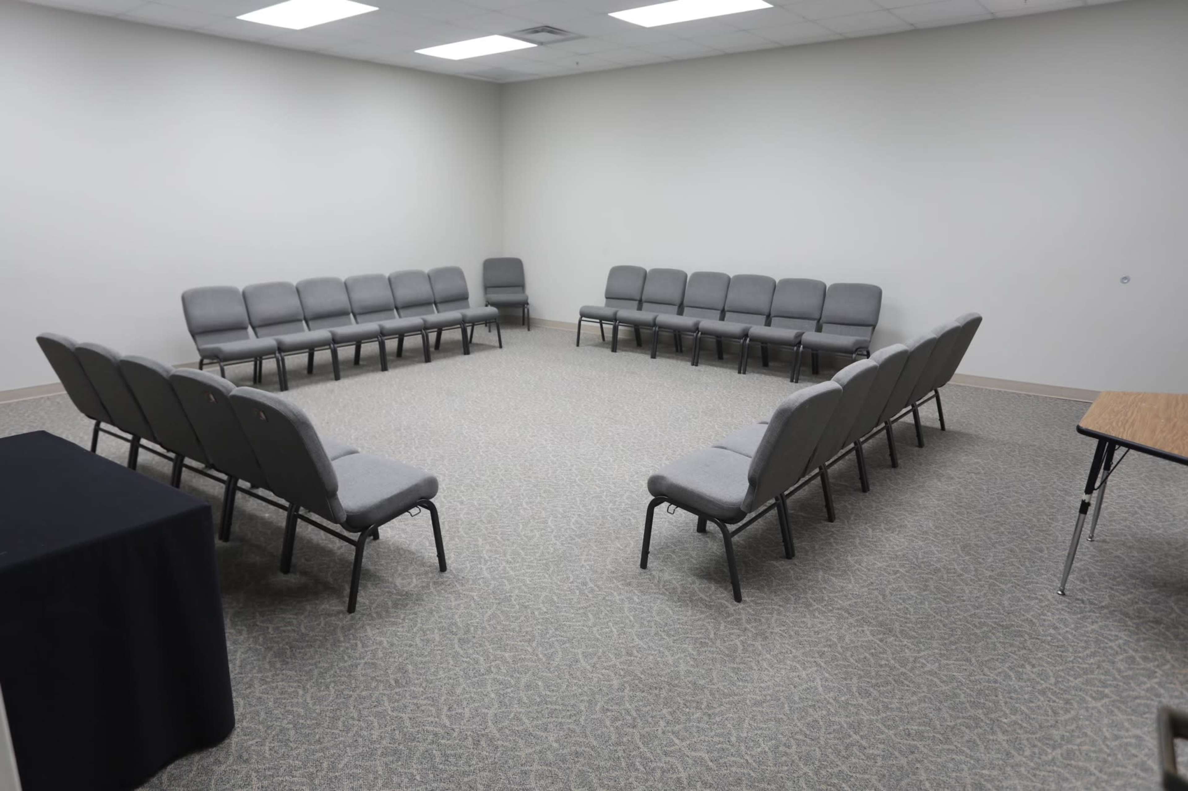 The image shows a well-lit room with a circular arrangement of gray chairs on a carpeted floor, with a table in the corner.