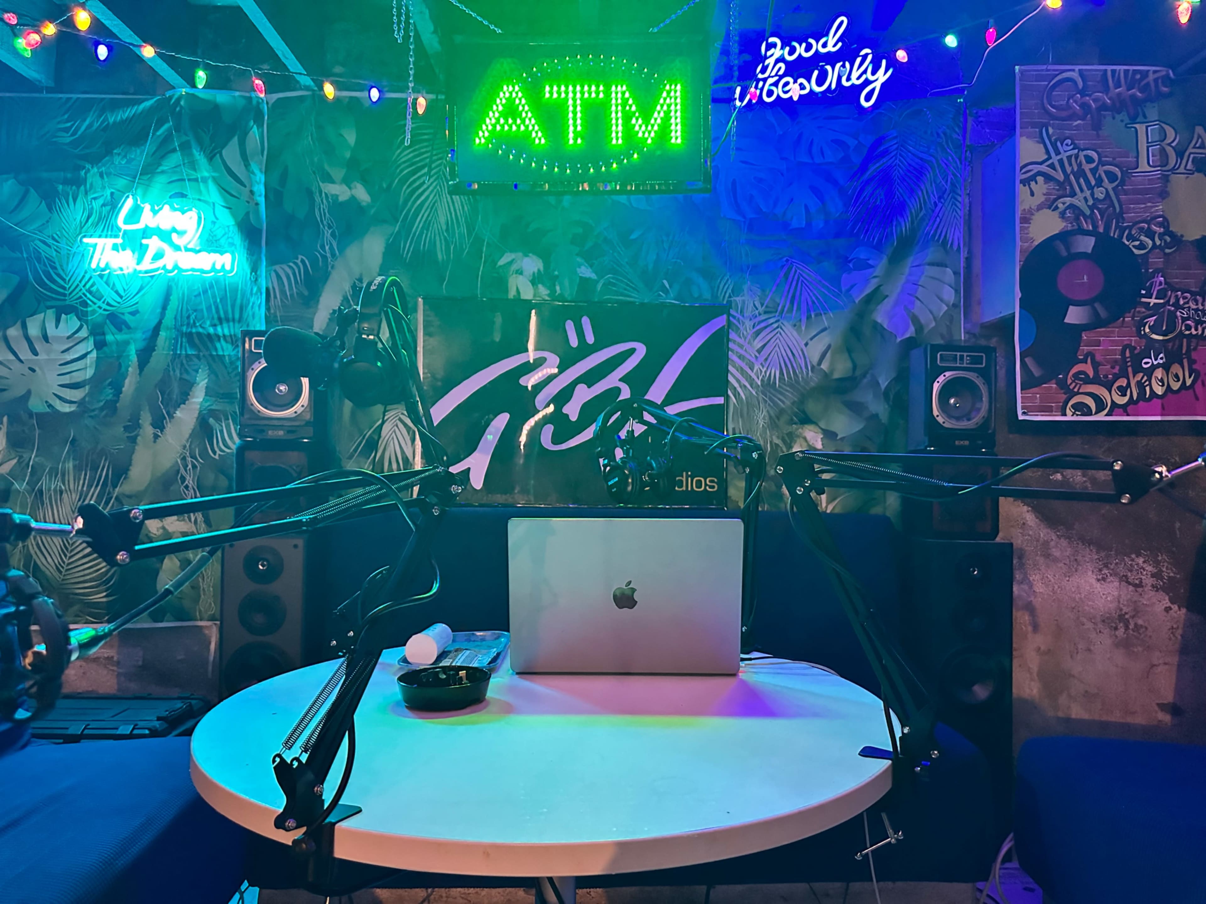A small studio setup features a table with a laptop, microphone arms, speakers, and colorful neon signs, all surrounded by a vibrant backdrop.