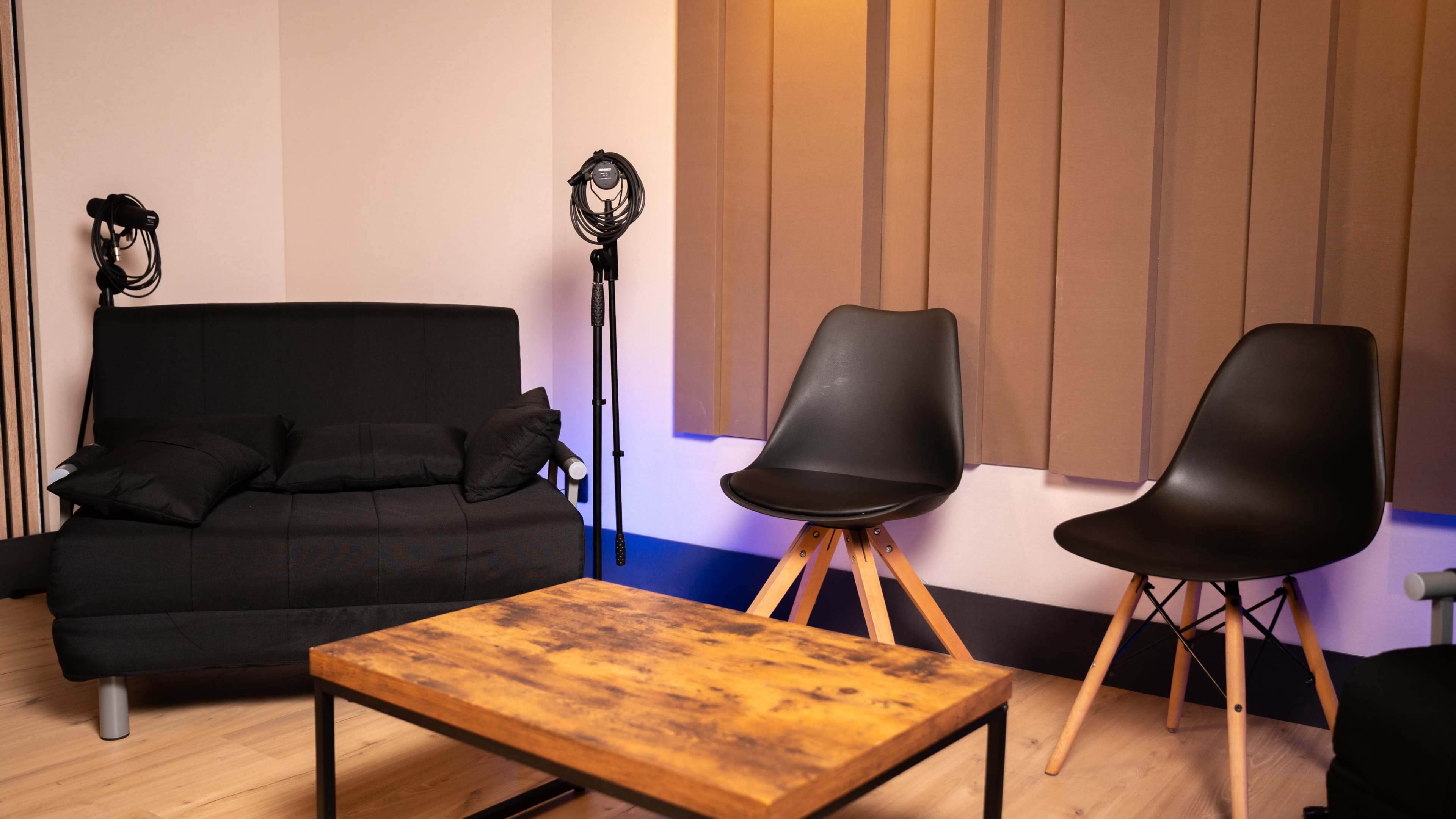 The image shows a small, modern room furnished with a dark sofa, two sleek chairs, and a wooden coffee table, with soft lighting and sound equipment visible in the corners.