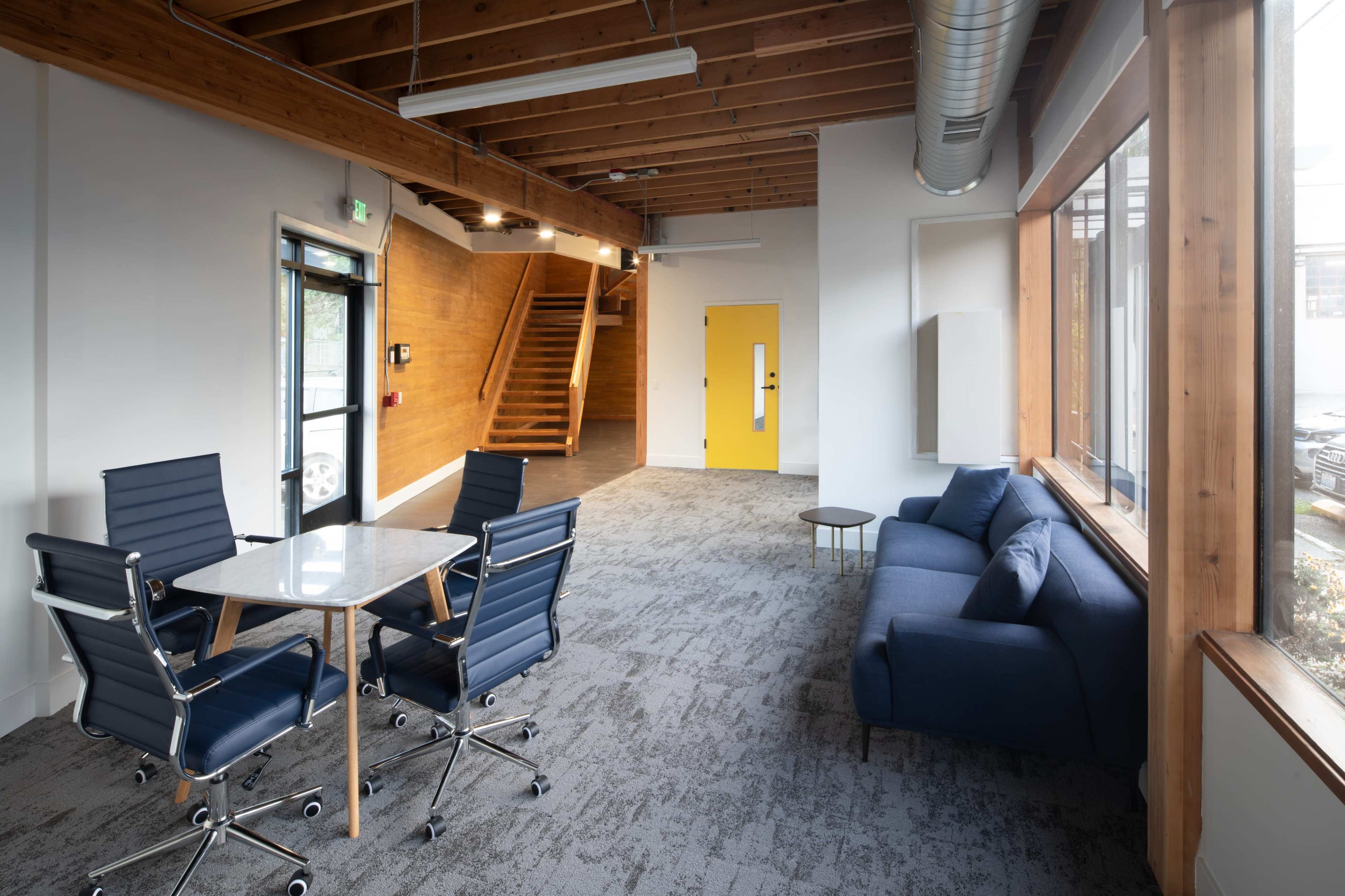 A modern office space featuring a small meeting area with a marble table and four chairs, a blue couch, wooden walls, and a staircase leading to an upper level.