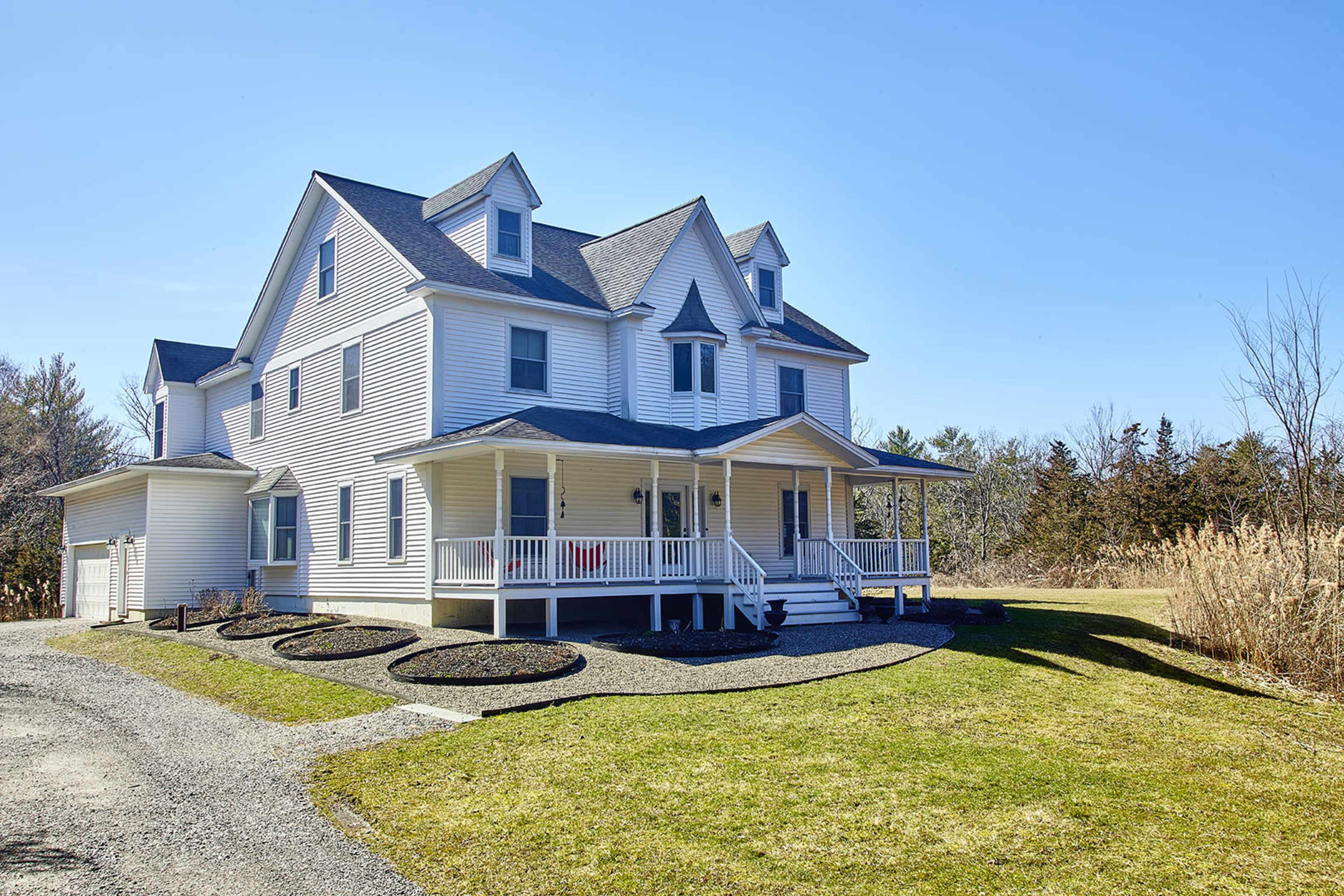 A large, white multi-story house with a wraparound porch is situated on a grassy plot beside a gravel driveway.