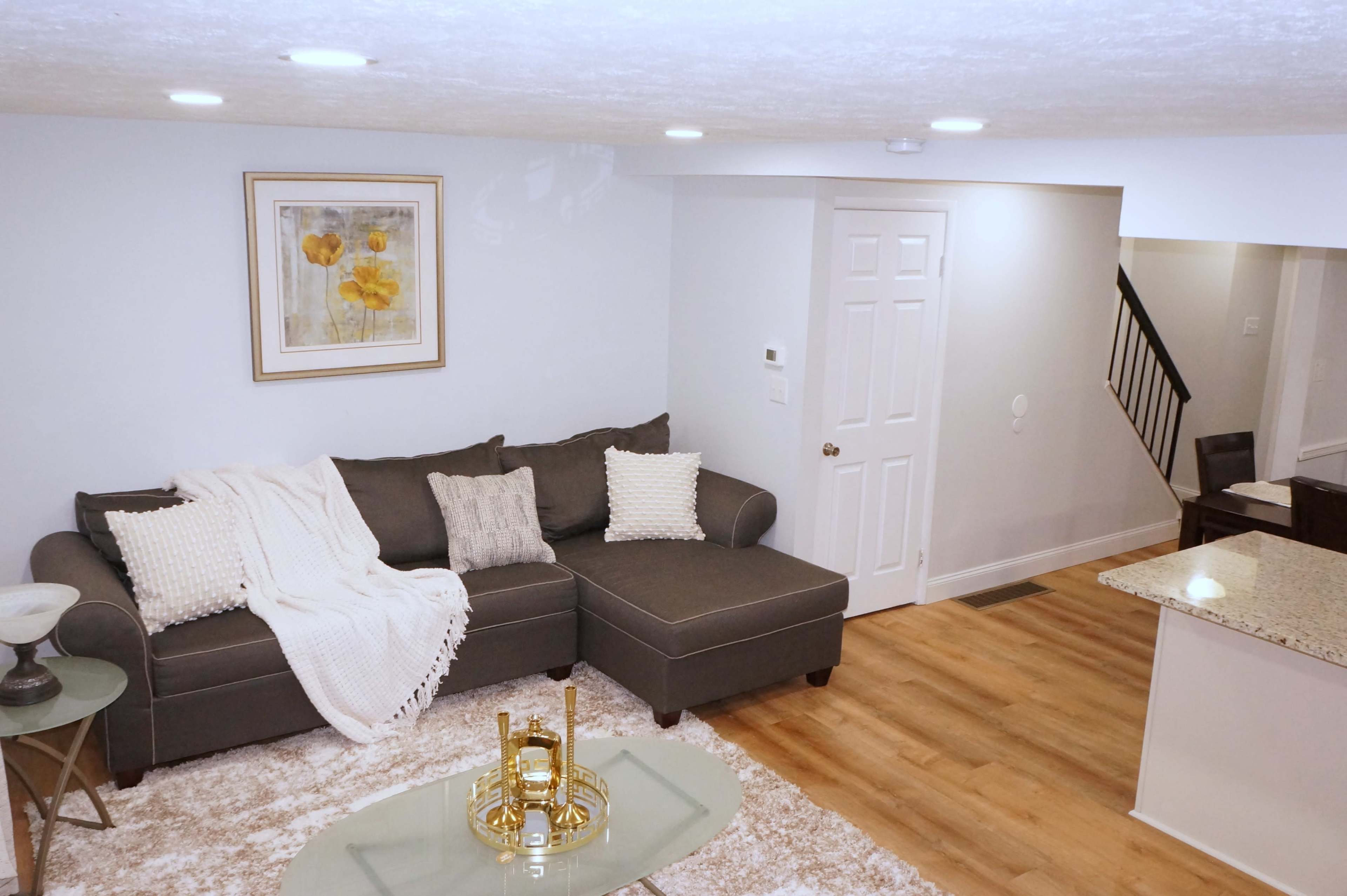 The image shows a modern living room with a gray sectional sofa, a glass coffee table, and a framed artwork on the wall, featuring neutral colors and a light wood floor.