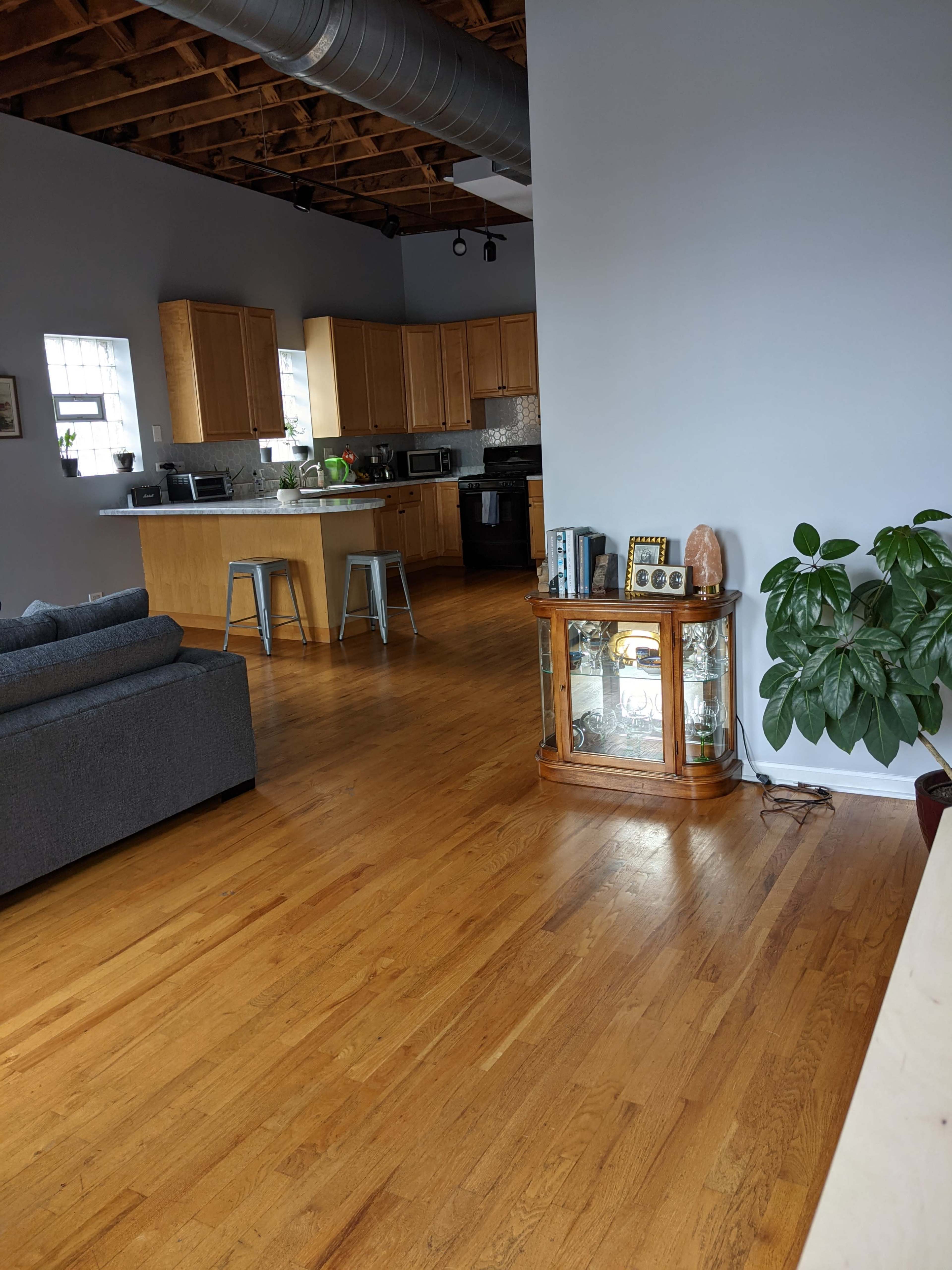 The image shows a spacious living area with wooden floors, a grey sofa, a glass cabinet displaying items, and a kitchen area in the background.
