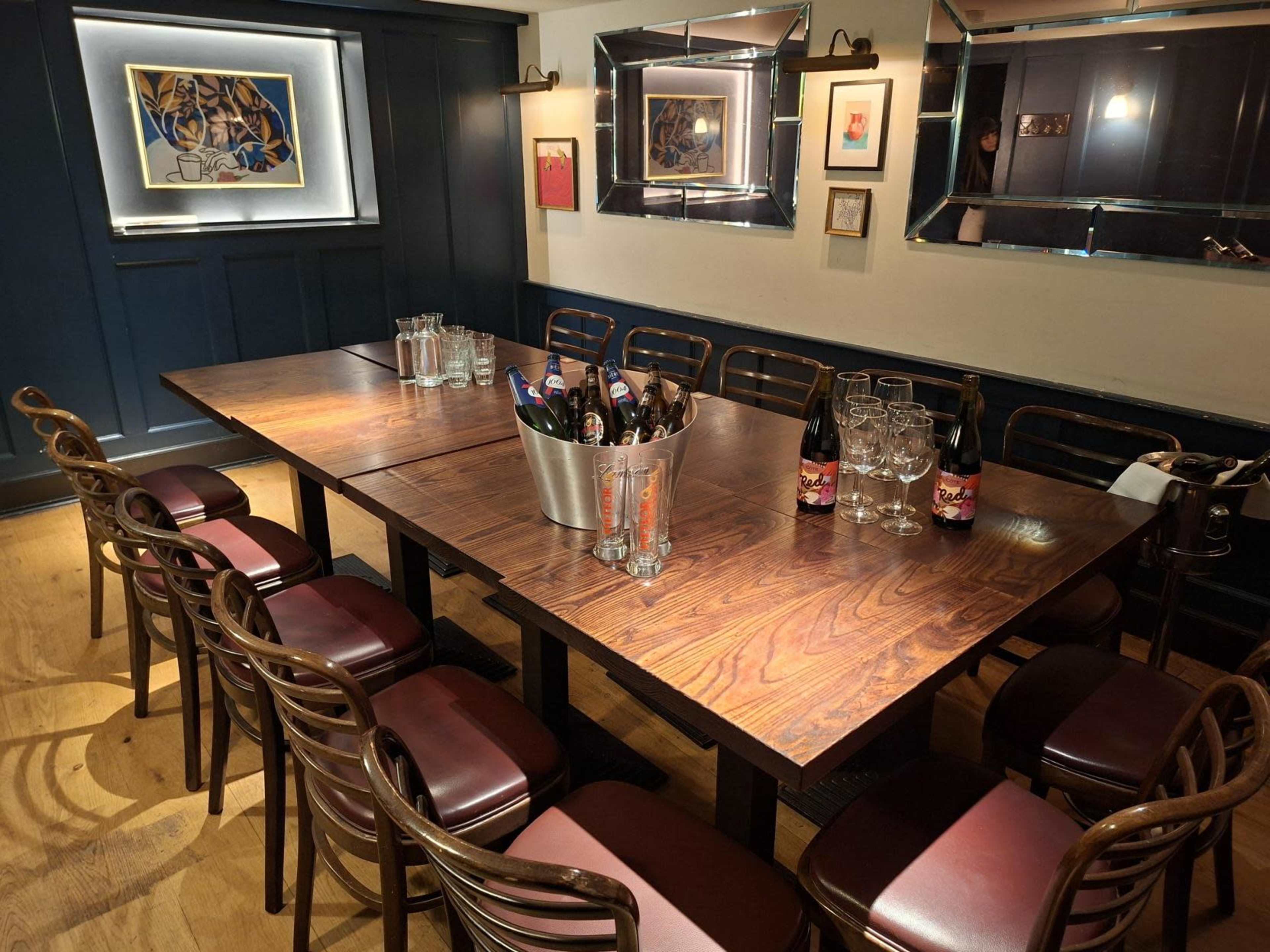 A long wooden table is set in a dimly lit room with brown chairs, featuring bottles of beer in an ice bucket and various artworks on the walls.