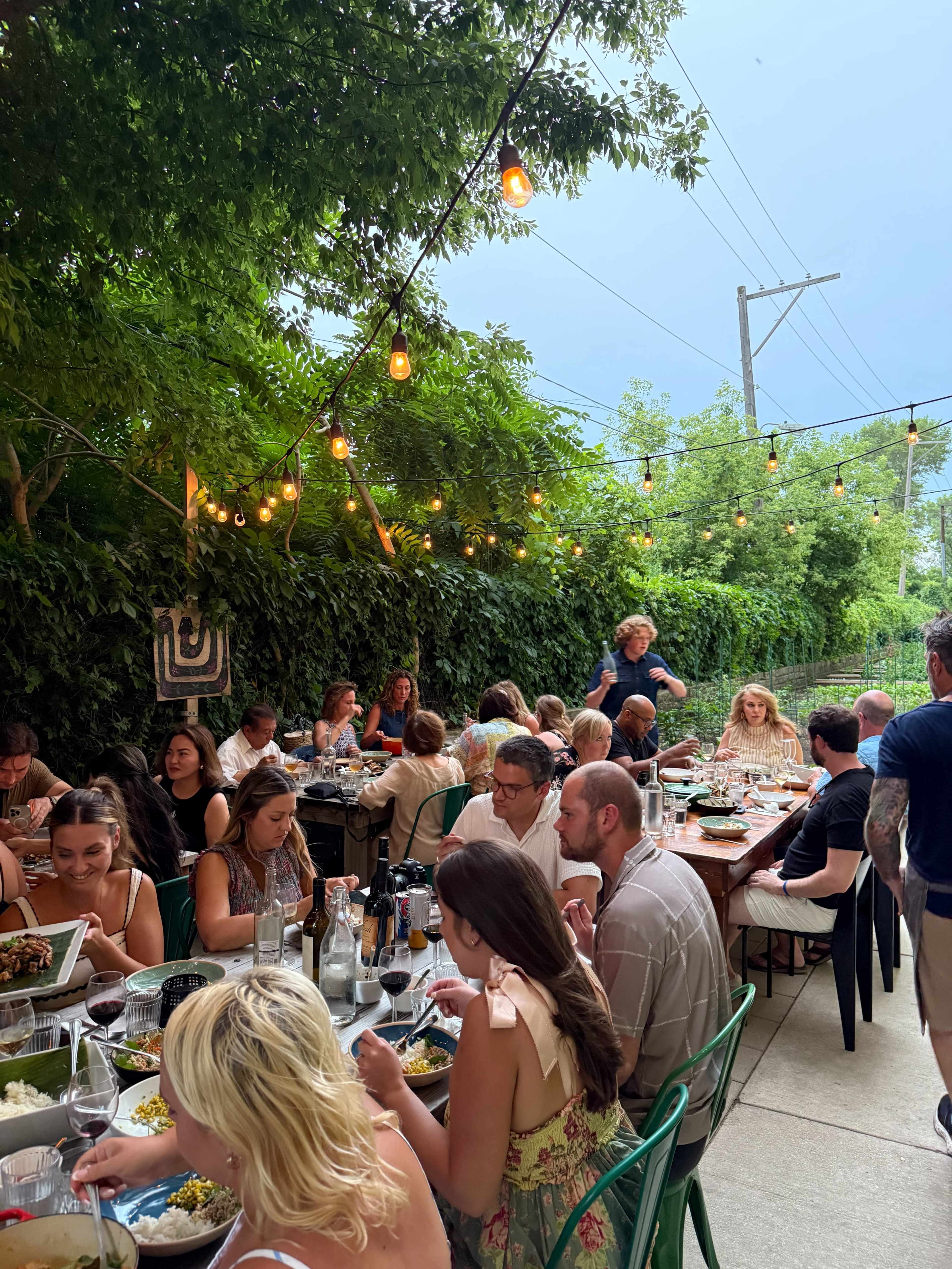 A group of people enjoy a meal at an outdoor dining area adorned with string lights and surrounded by greenery.
