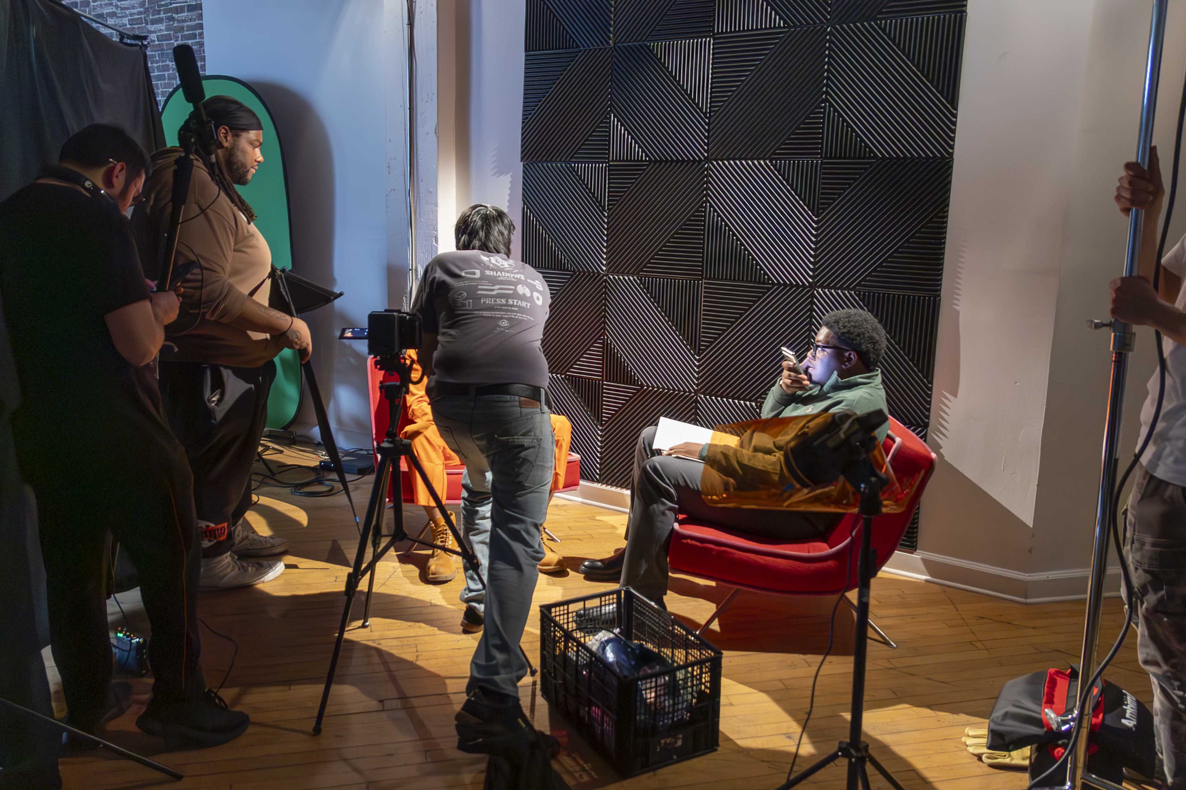 A group of people are working in a studio setting, setting up equipment around a man seated in a red chair who is reading a script.