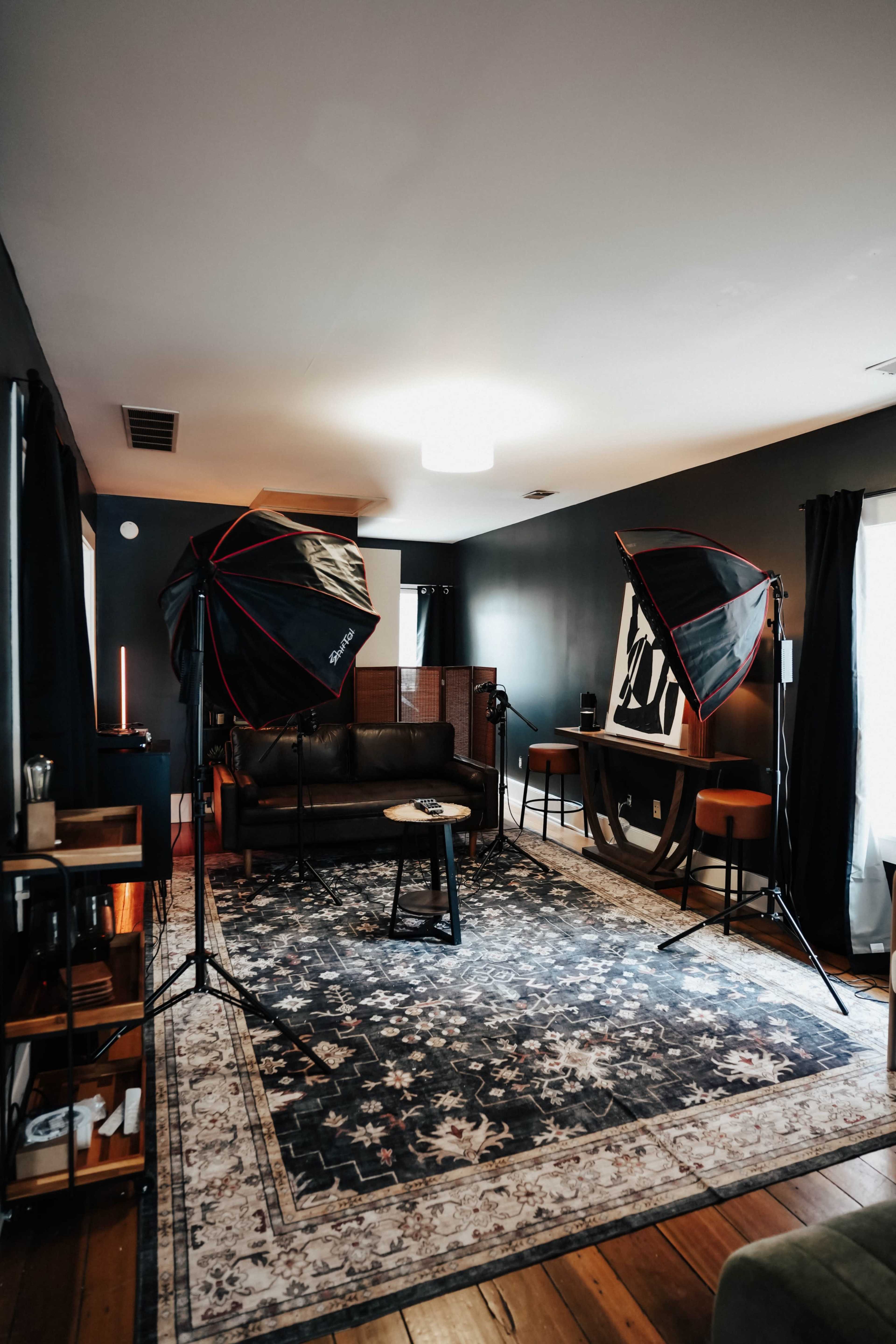 The image shows a photography studio setup featuring softbox lights, a dark-colored couch, and a patterned rug on a wooden floor.