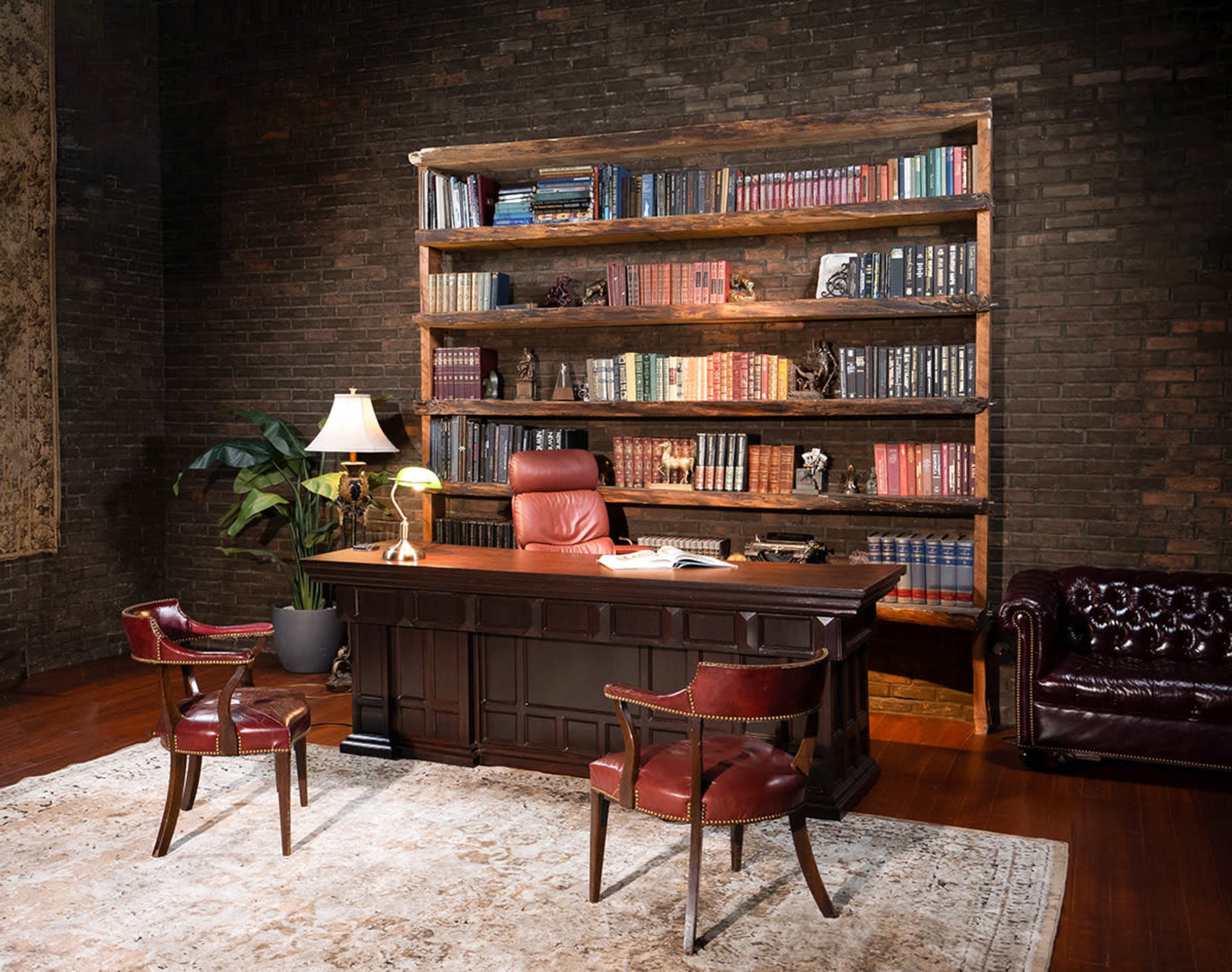 A wooden desk with a red chair is positioned in front of a bookshelf filled with books, surrounded by a dark brick wall and a plant, with a lamp providing light.