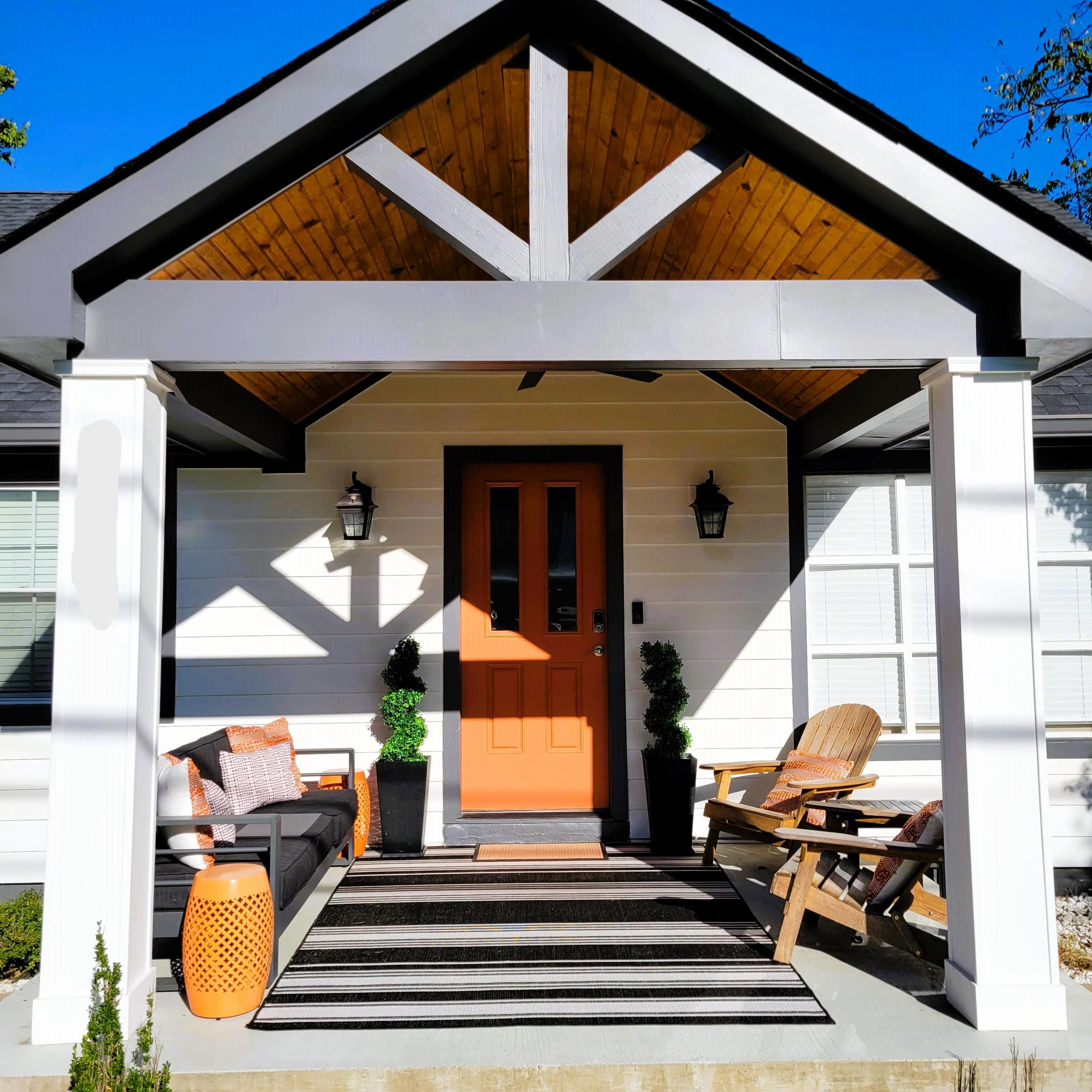 The image shows a front porch with an orange door, decorative seating, and potted plants underneath a wooden ceiling.
