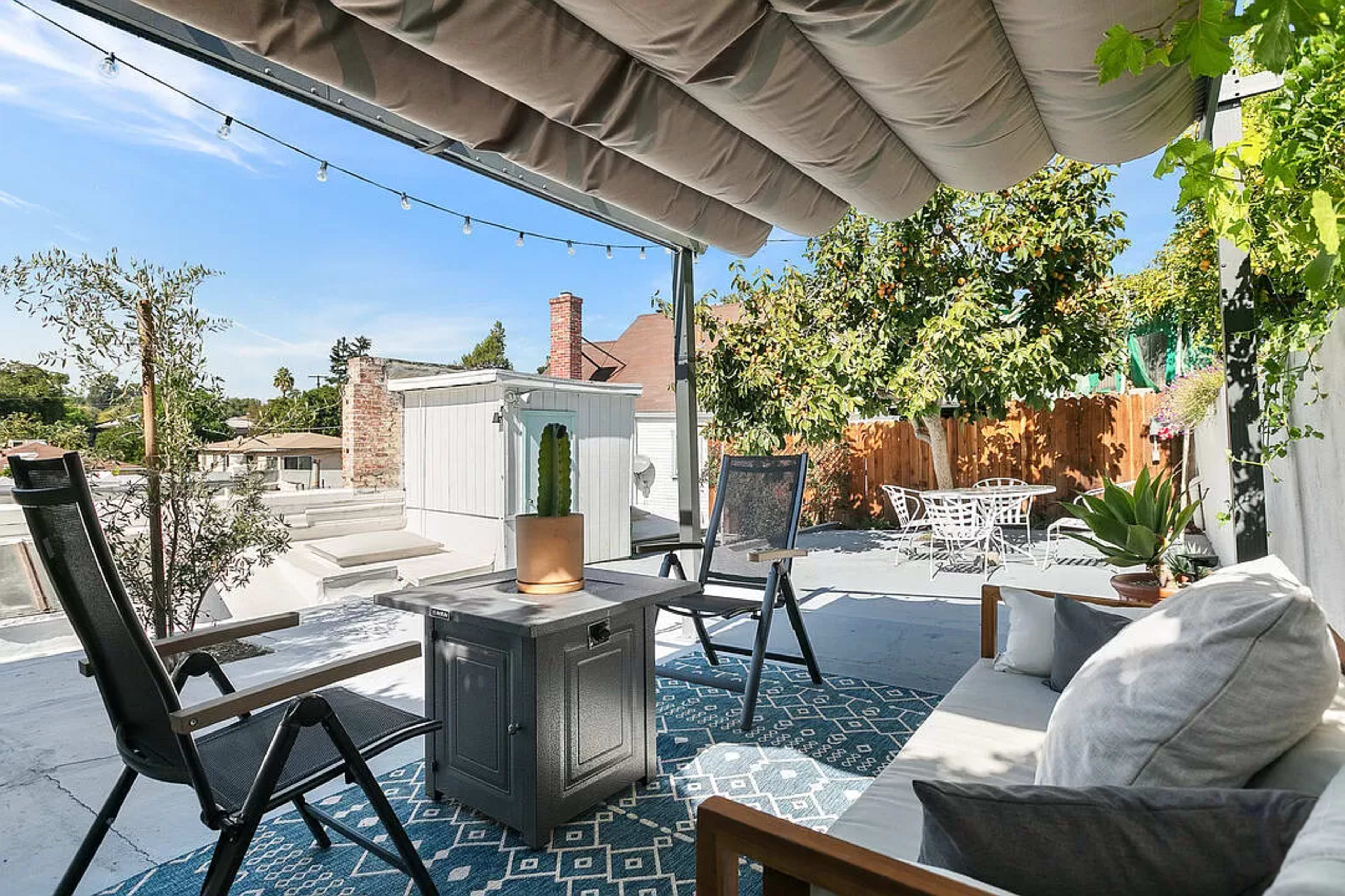 The image shows a rooftop patio with outdoor seating, a small table, and potted plants, surrounded by greenery and string lights overhead.