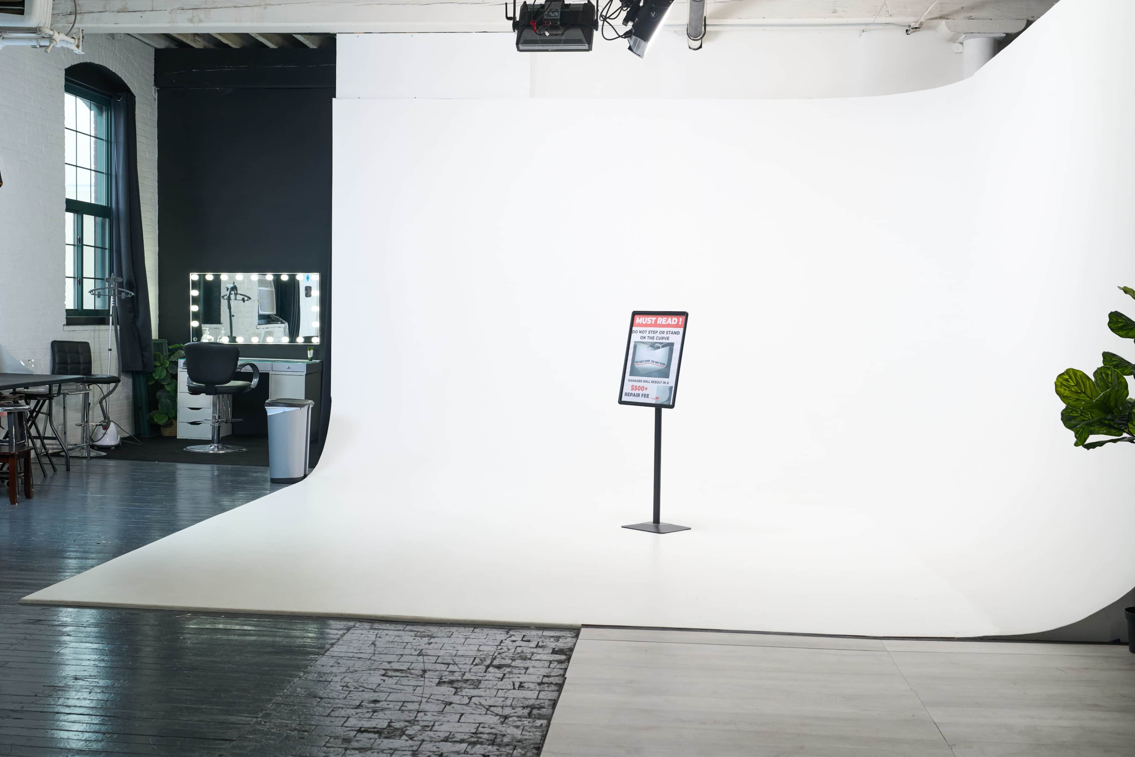 The image shows a photography studio with a large white backdrop, a sign on a stand in the foreground, and a makeup station with a mirror and chair in the background.