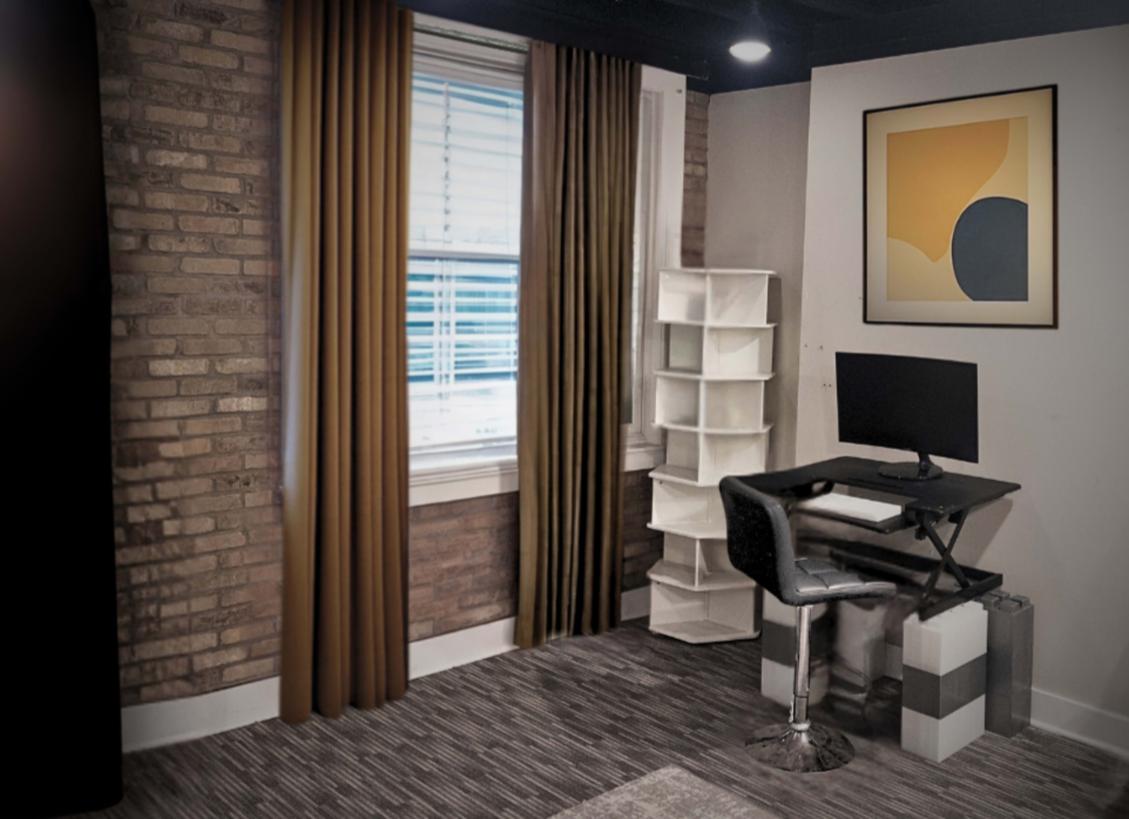 The image shows a modern workspace featuring a desk with a computer, a chair, and a window with brown curtains, alongside a white shelving unit and a graphic art piece on the wall.