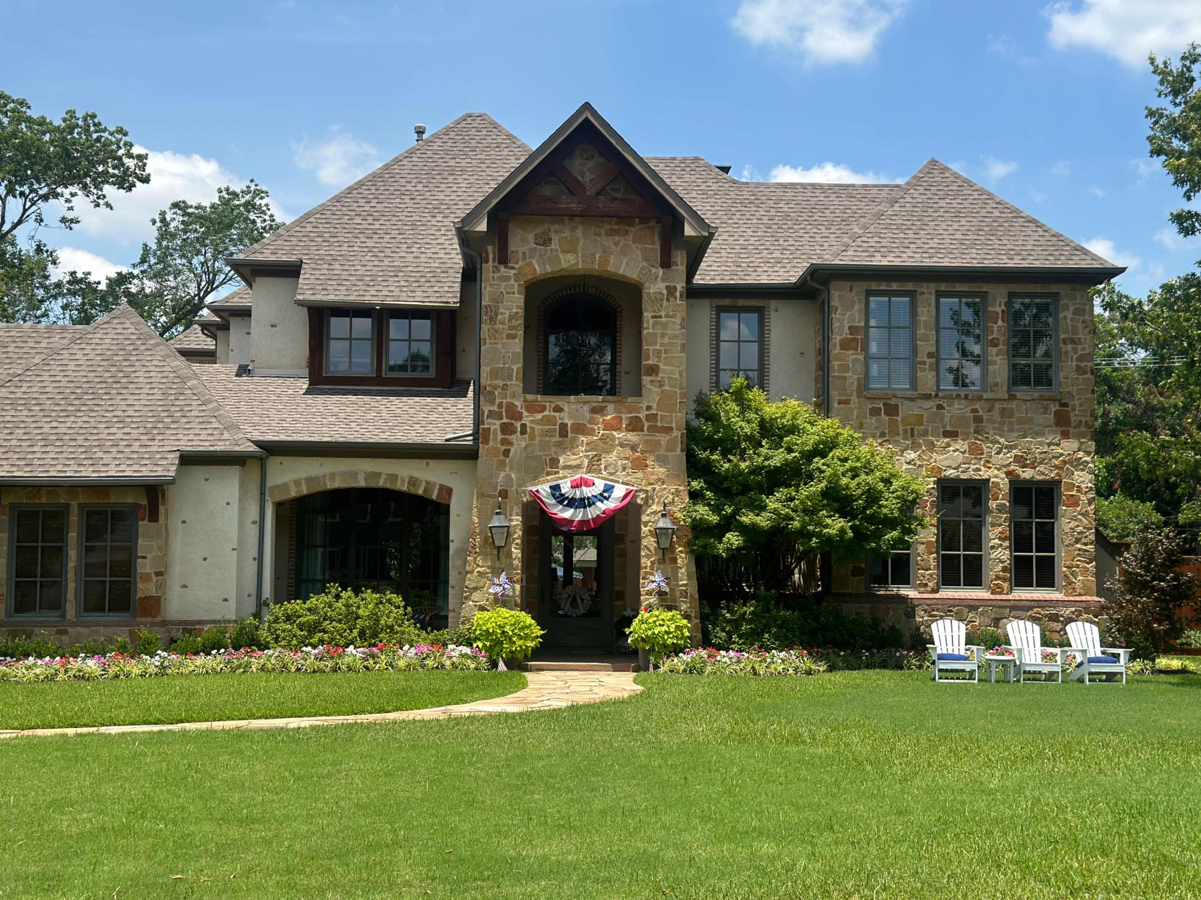 A large, two-story stone and stucco house with a landscaped front yard featuring flower beds and white adirondack chairs.