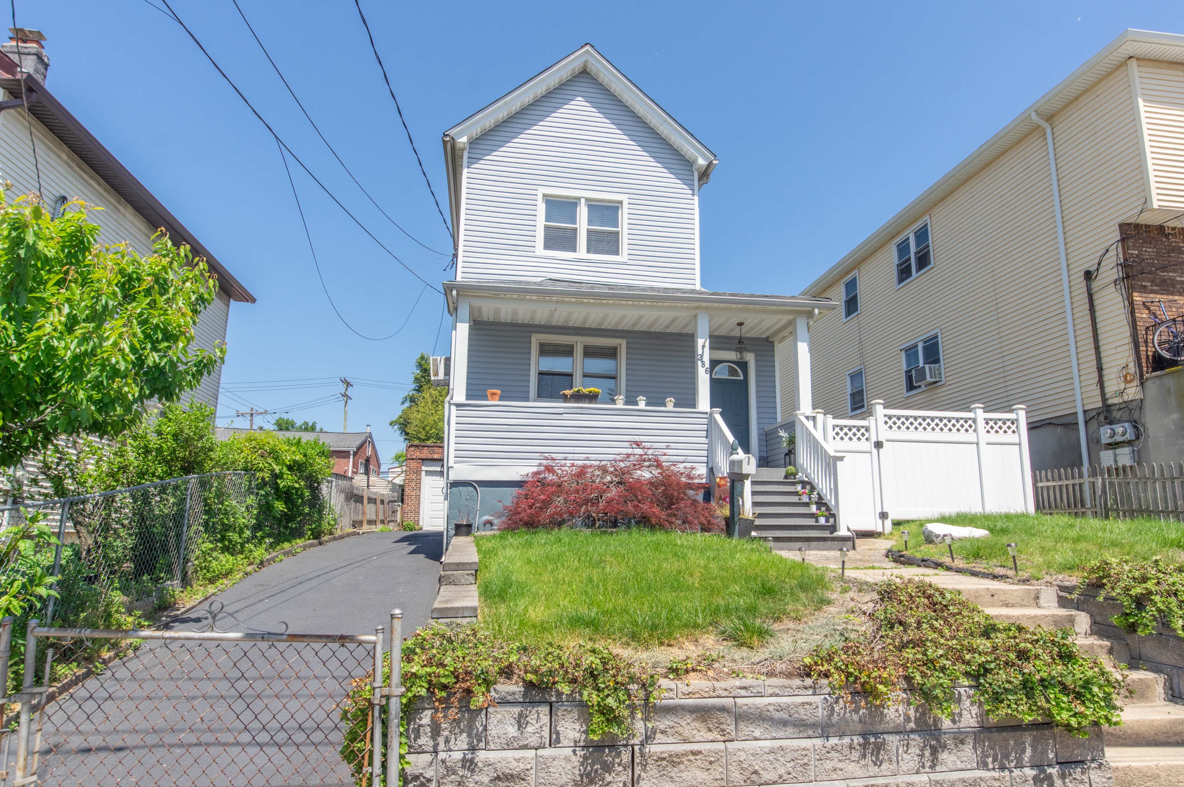 A two-story blue house with a front porch is situated on a grassy lot, bordered by a sidewalk and a fence.