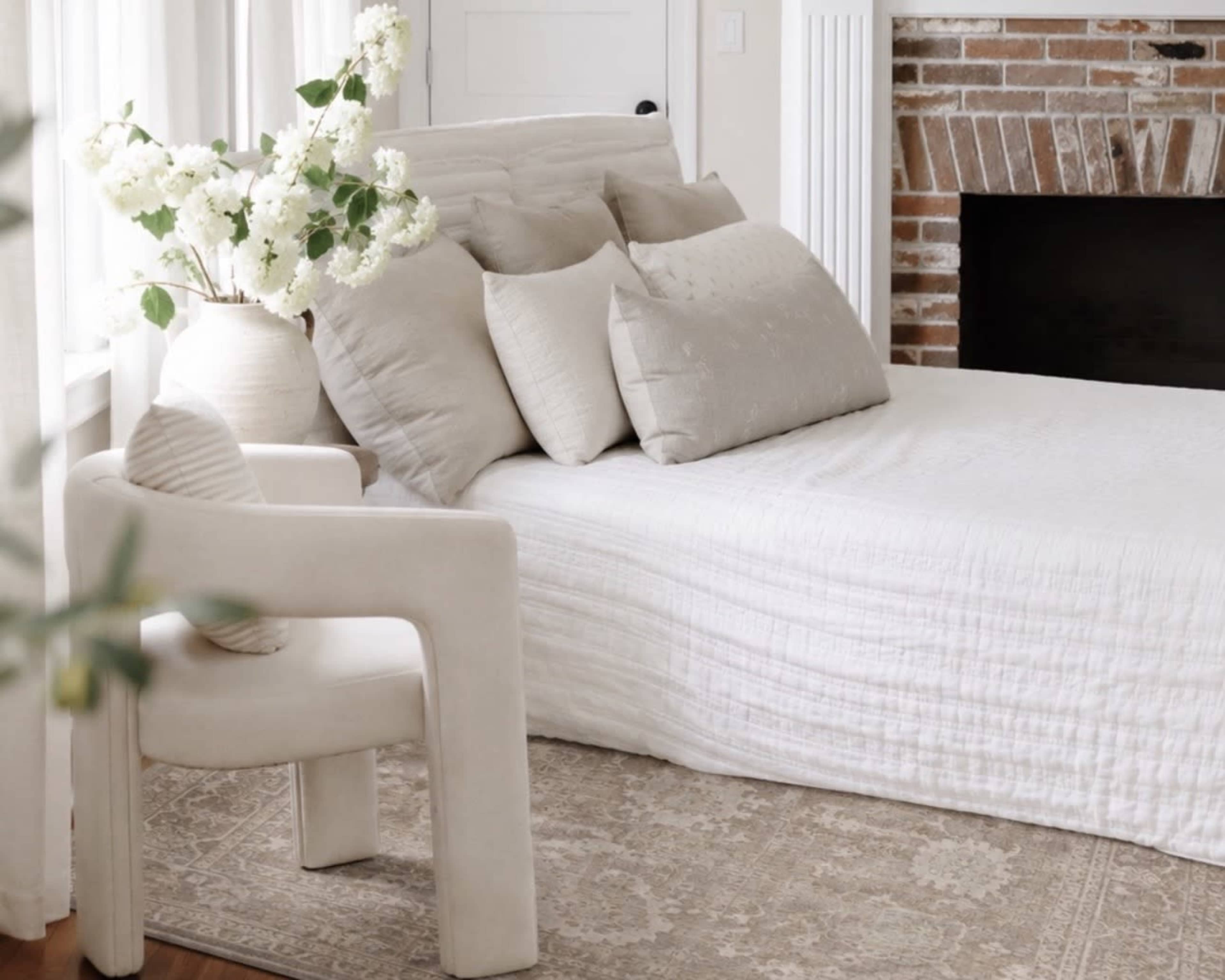 The image features a cozy bedroom with a bed adorned in light linens, several decorative pillows, a minimalist chair, and a vase of white flowers beside a brick fireplace.