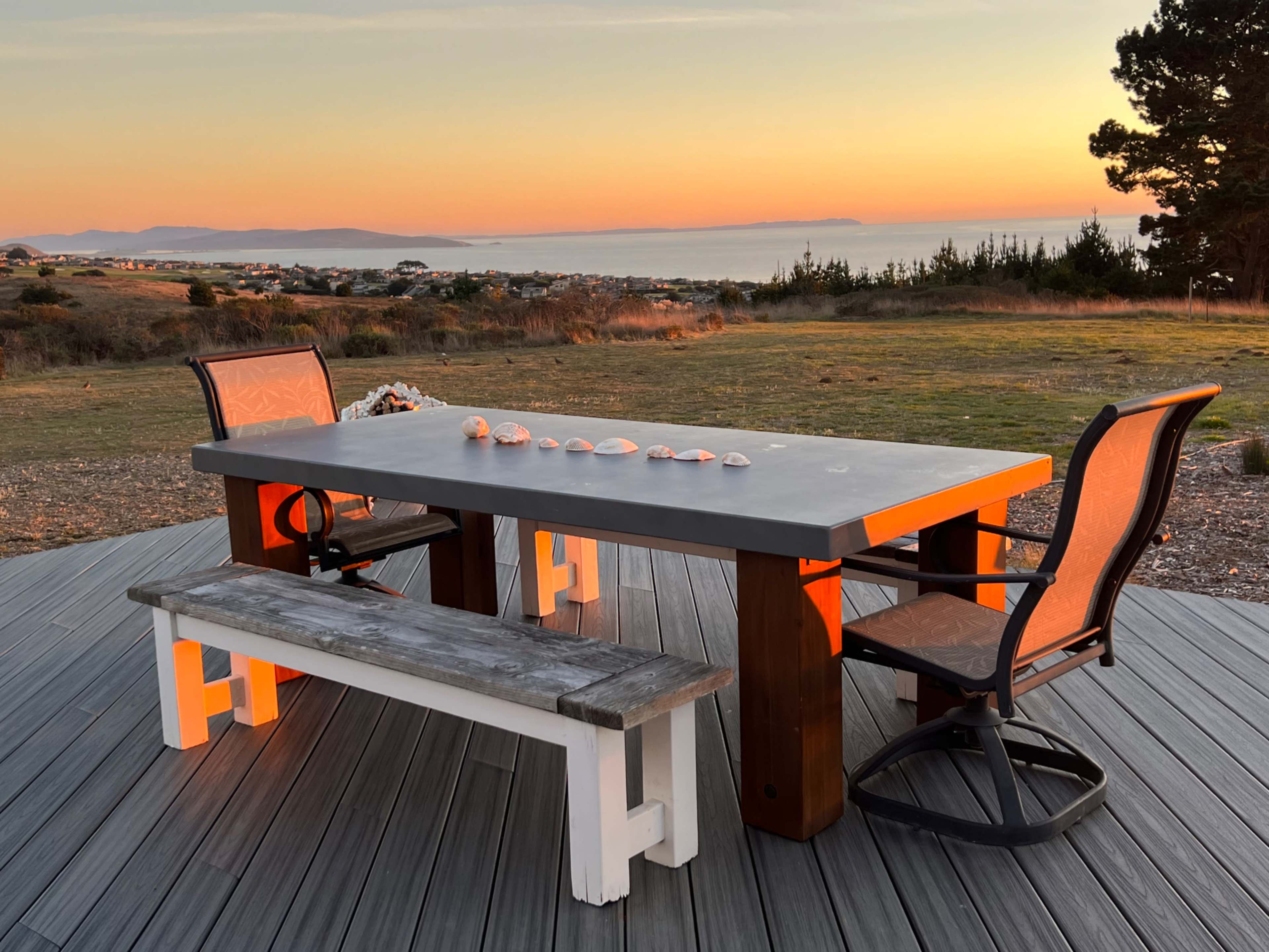 A concrete table and two chairs are set against a backdrop of a sunset over the ocean, with a sandy area and distant hills visible.