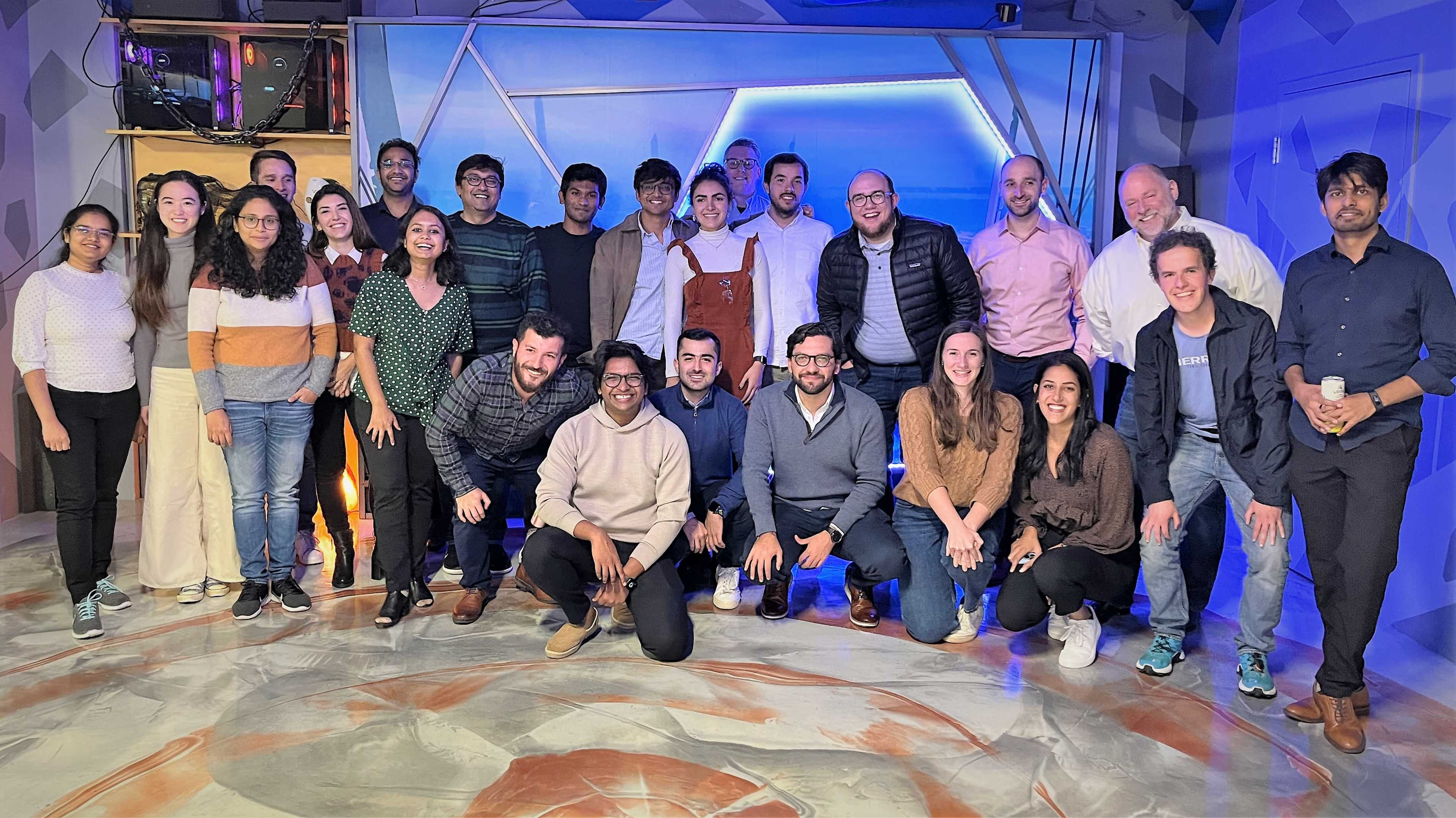 A diverse group of approximately thirty people poses together in a modern studio setting with blue lighting.