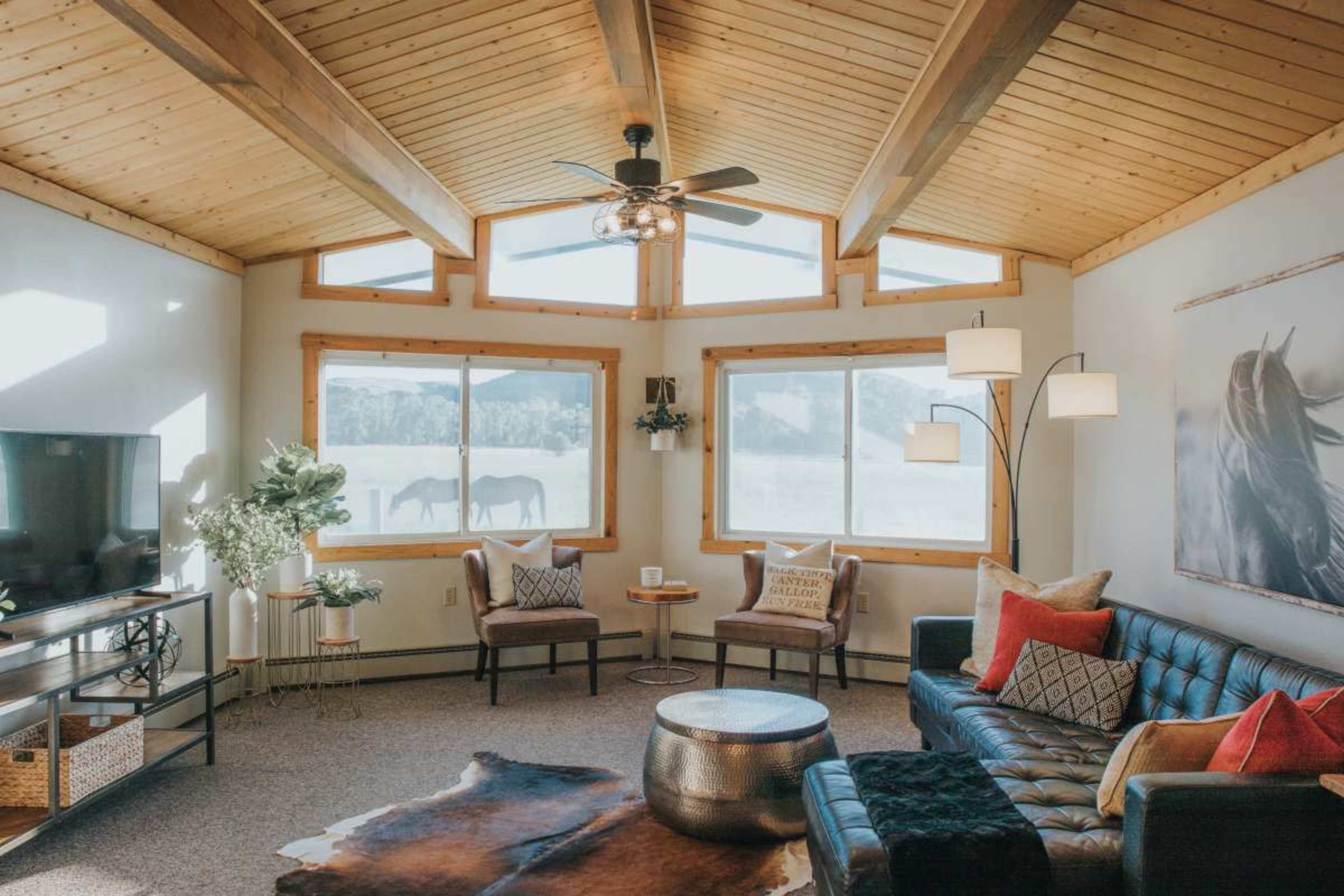 The image shows a modern living room with a wood-beamed ceiling, large windows, a leather sofa, two chairs, and a decorative coffee table, with horses visible outside.