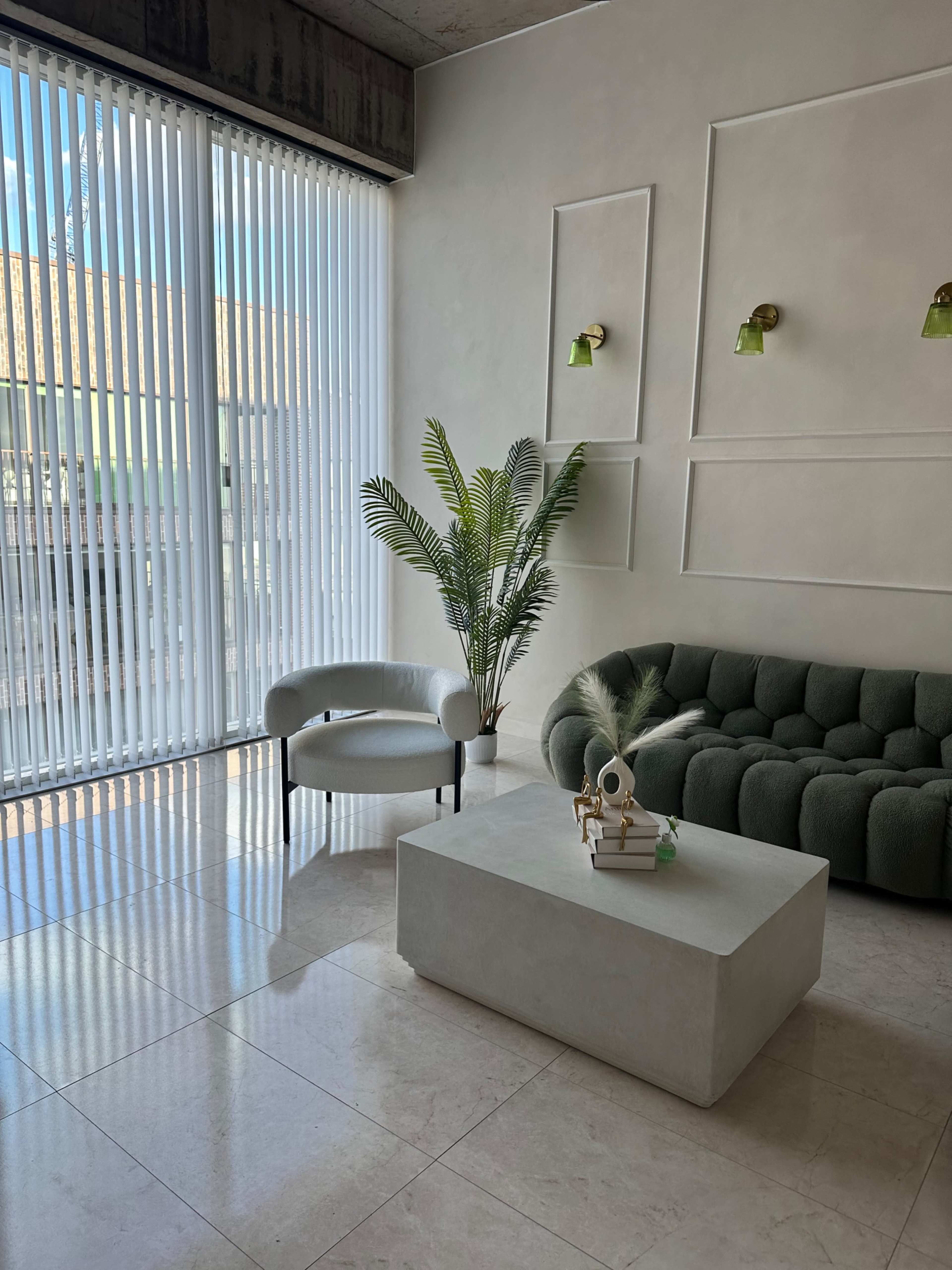 The image features a modern living room with a green couch, a white coffee table, a potted plant, and large windows covered with vertical blinds.