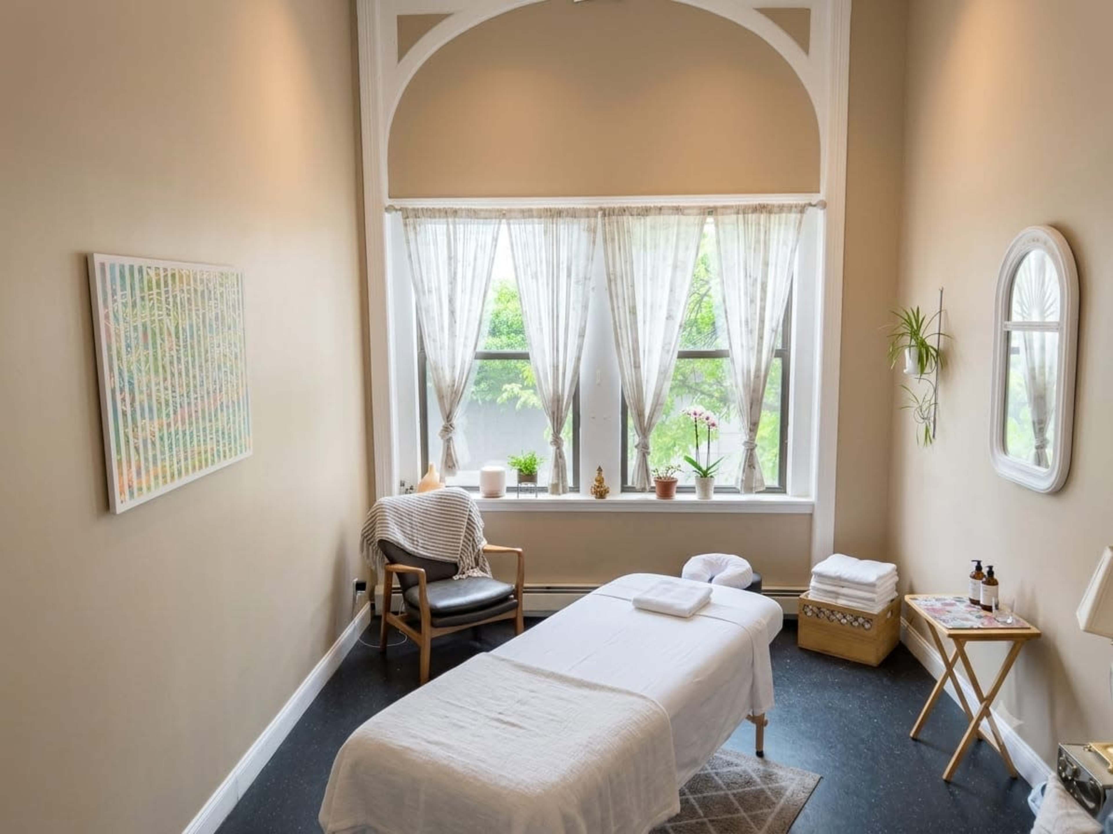 The image shows a serene massage therapy room with a treatment table, a chair, and large windows adorned with sheer curtains.