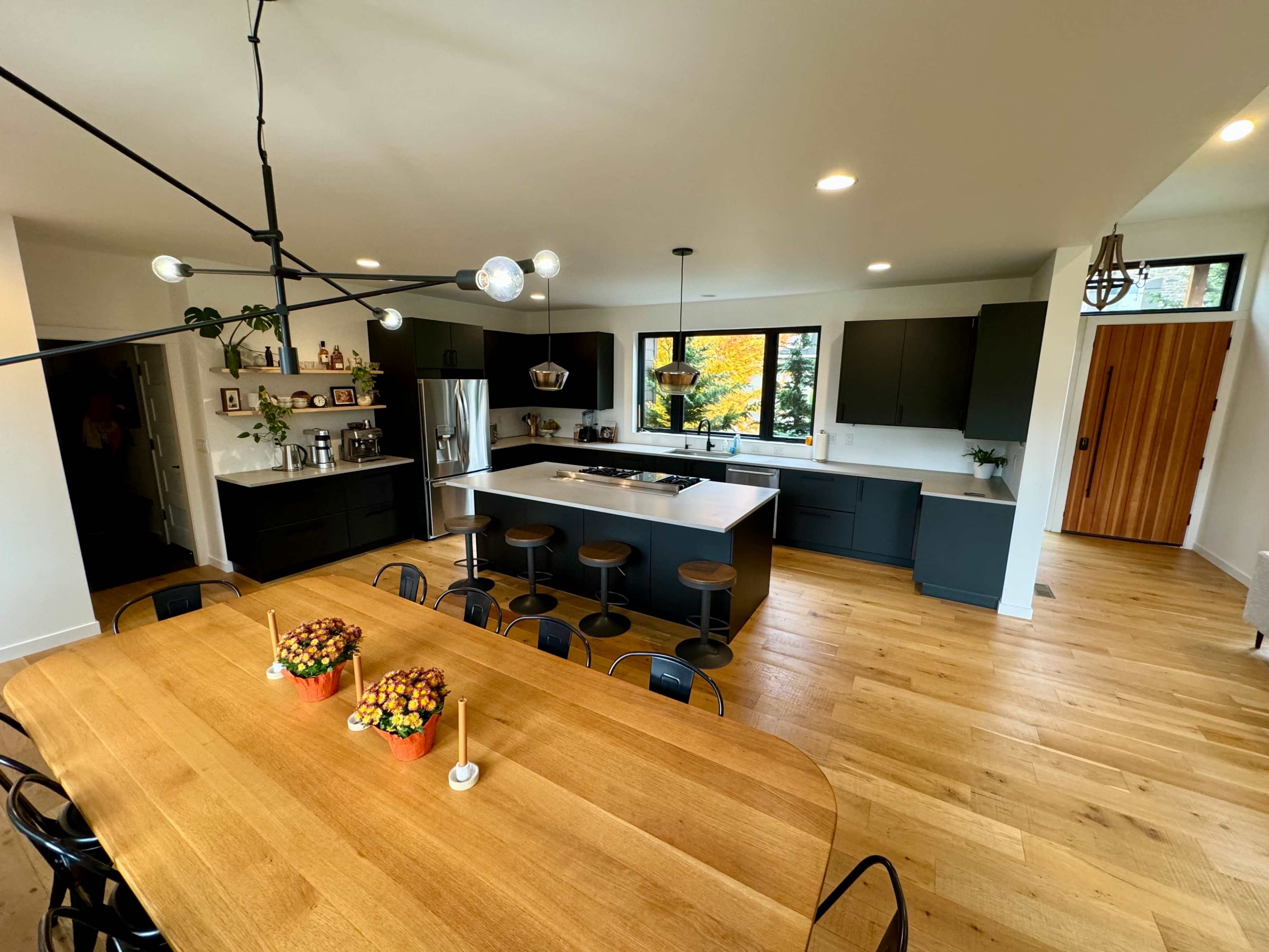 A modern kitchen and dining area featuring a large wooden table, black cabinets, a white countertop, and a bright window with autumn foliage visible outside.