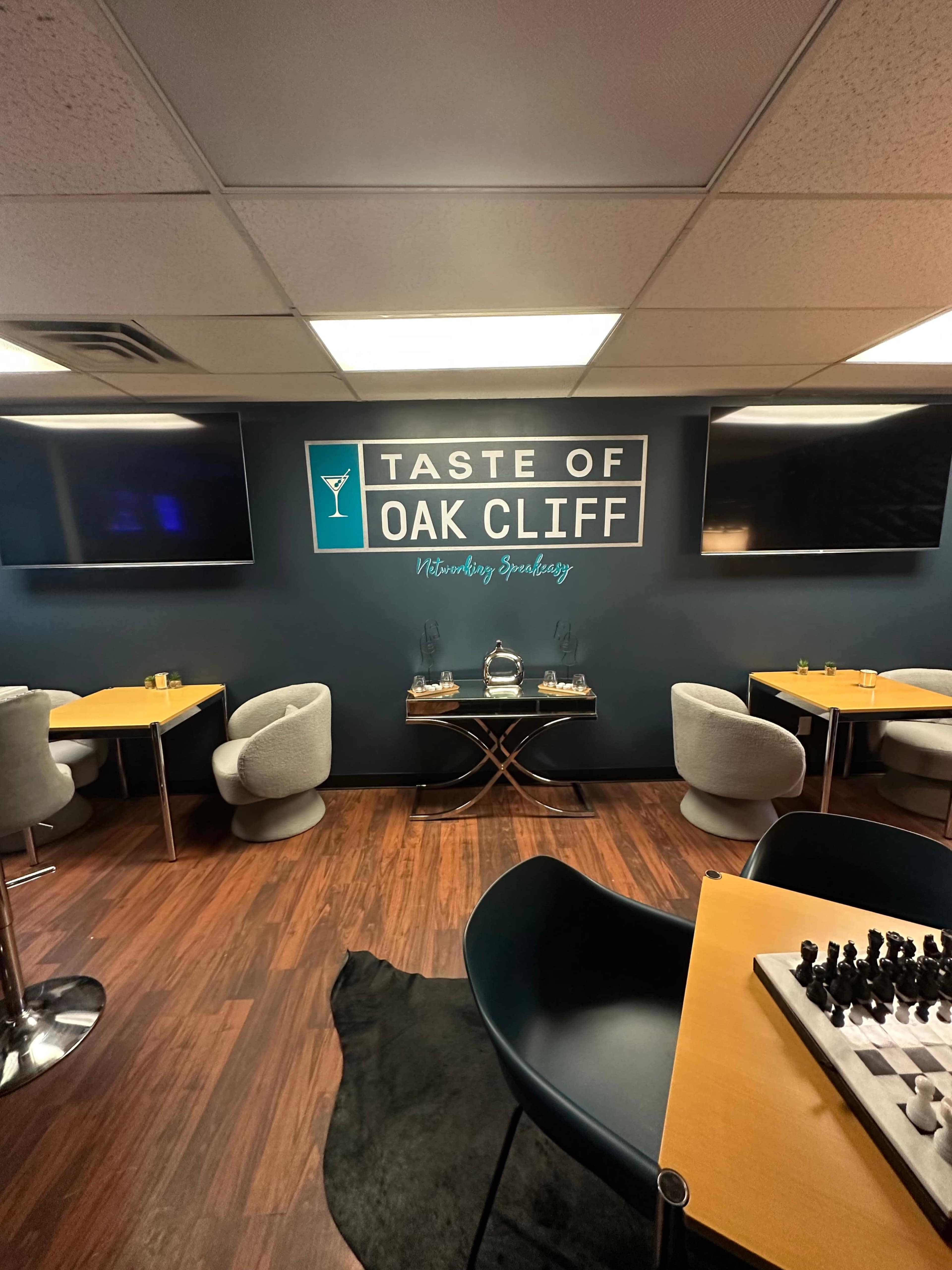 The image shows a modern lounge setting with several tables, plush chairs, two large wall-mounted TVs, and a chessboard on a table.