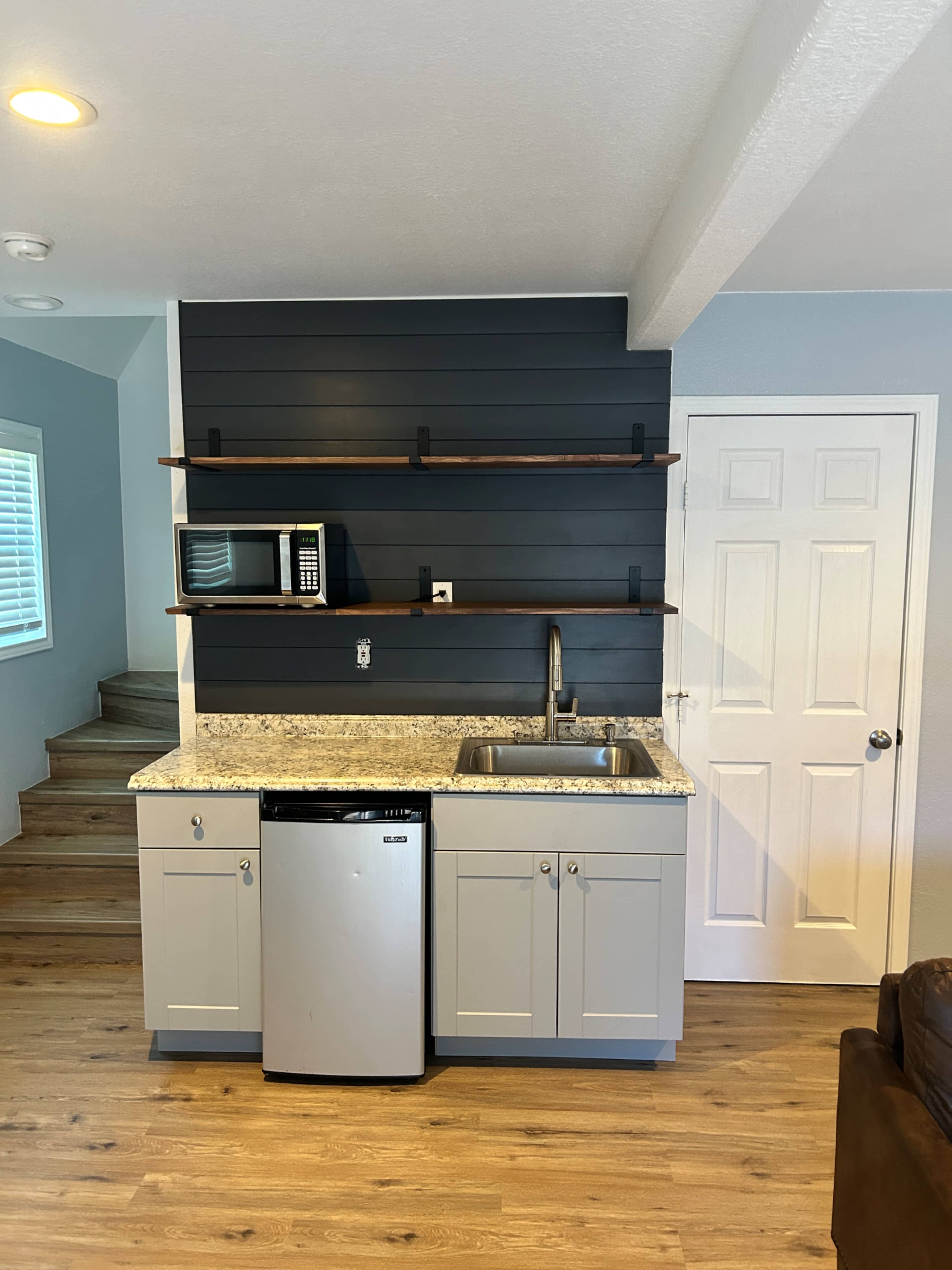A compact kitchen area with gray cabinets, a stainless steel refrigerator, a sink, a microwave, and wooden shelves against a dark wall.