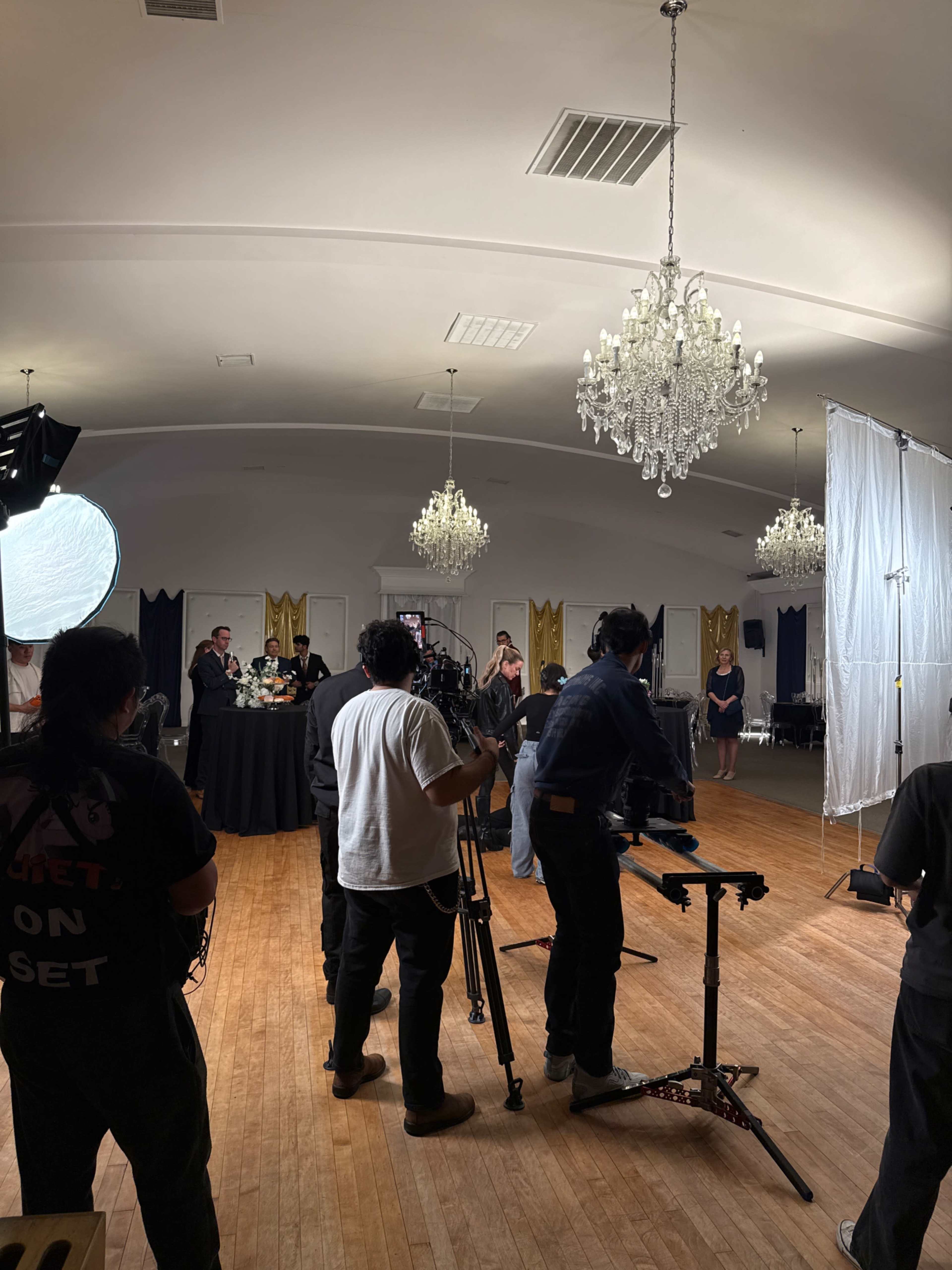A film crew is set up in a spacious room with chandeliers, preparing to shoot a scene involving a group of people around a table.