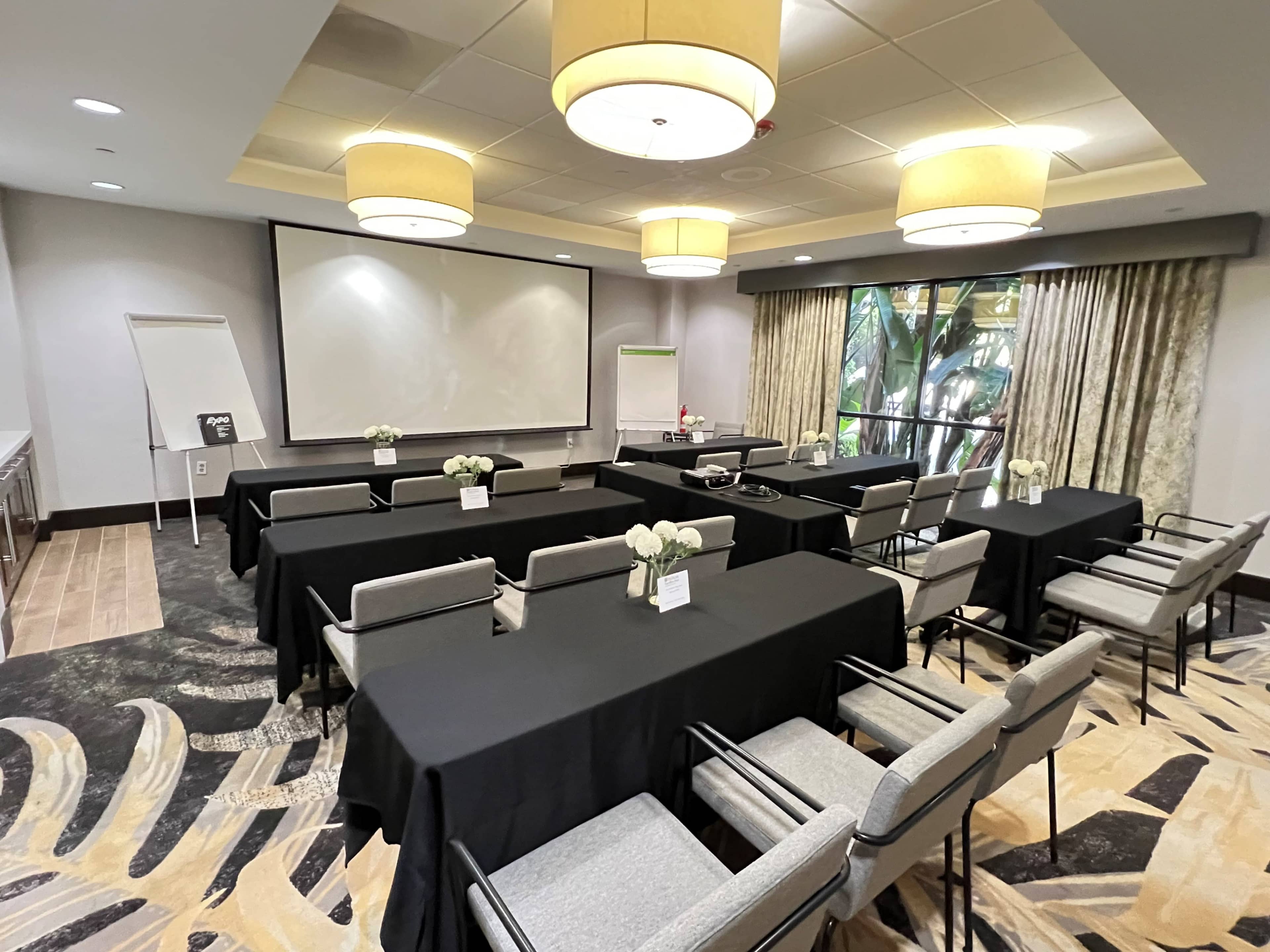A conference room is set up with several black-covered tables, chairs, and a large screen at the front, with natural light coming through large windows.