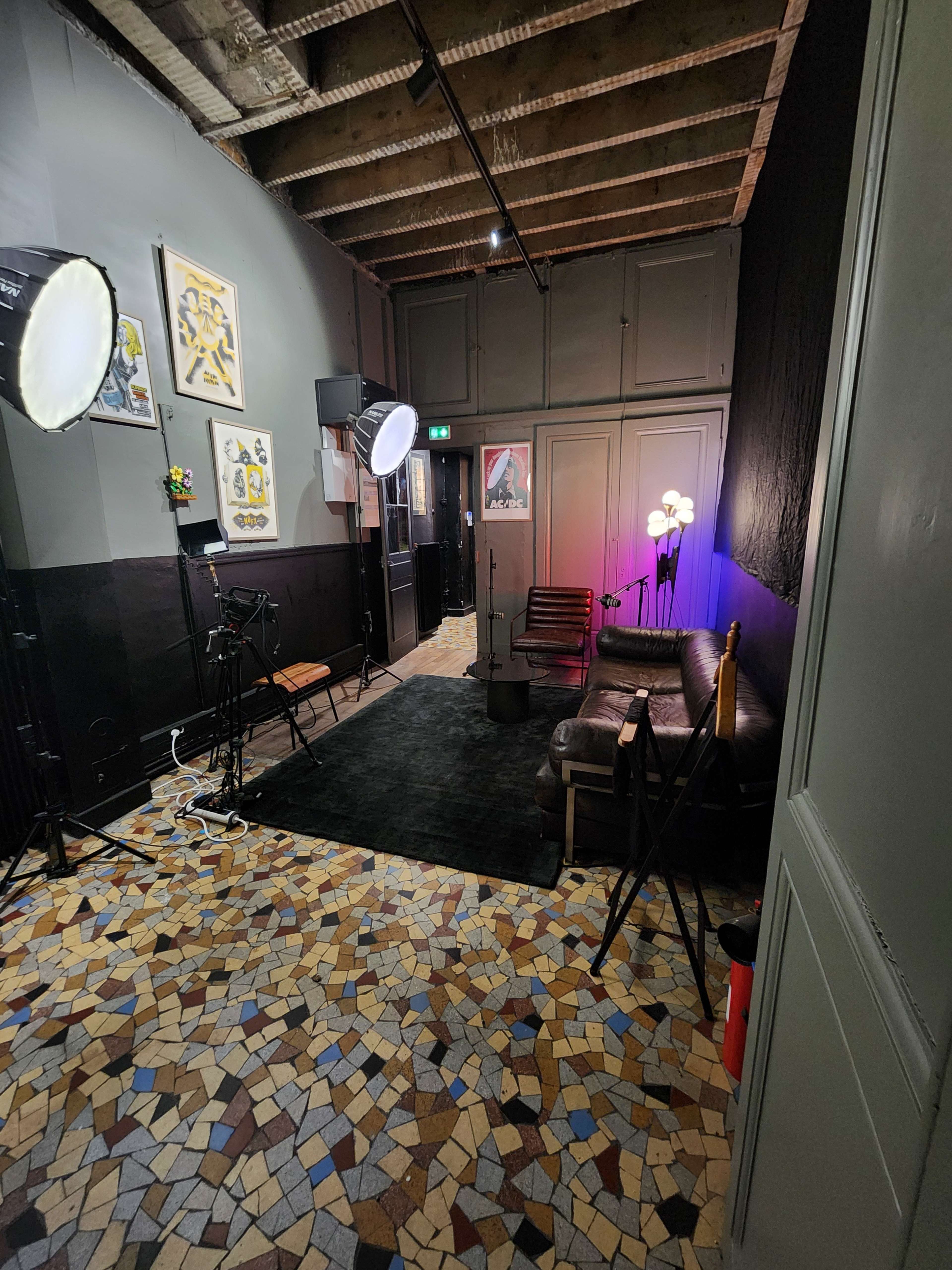 A small, dimly lit room with a patterned tiled floor, featuring a couch, lighting equipment, and artwork on the walls.
