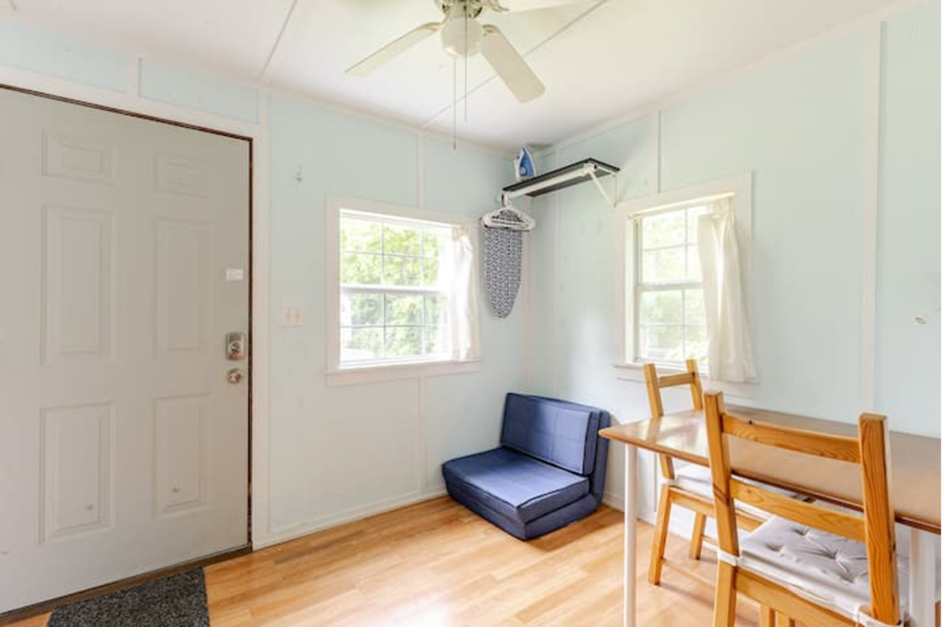 The room features a light blue wall, a front door, a small seating area with a blue couch, a table with wooden chairs, and two windows allowing natural light.