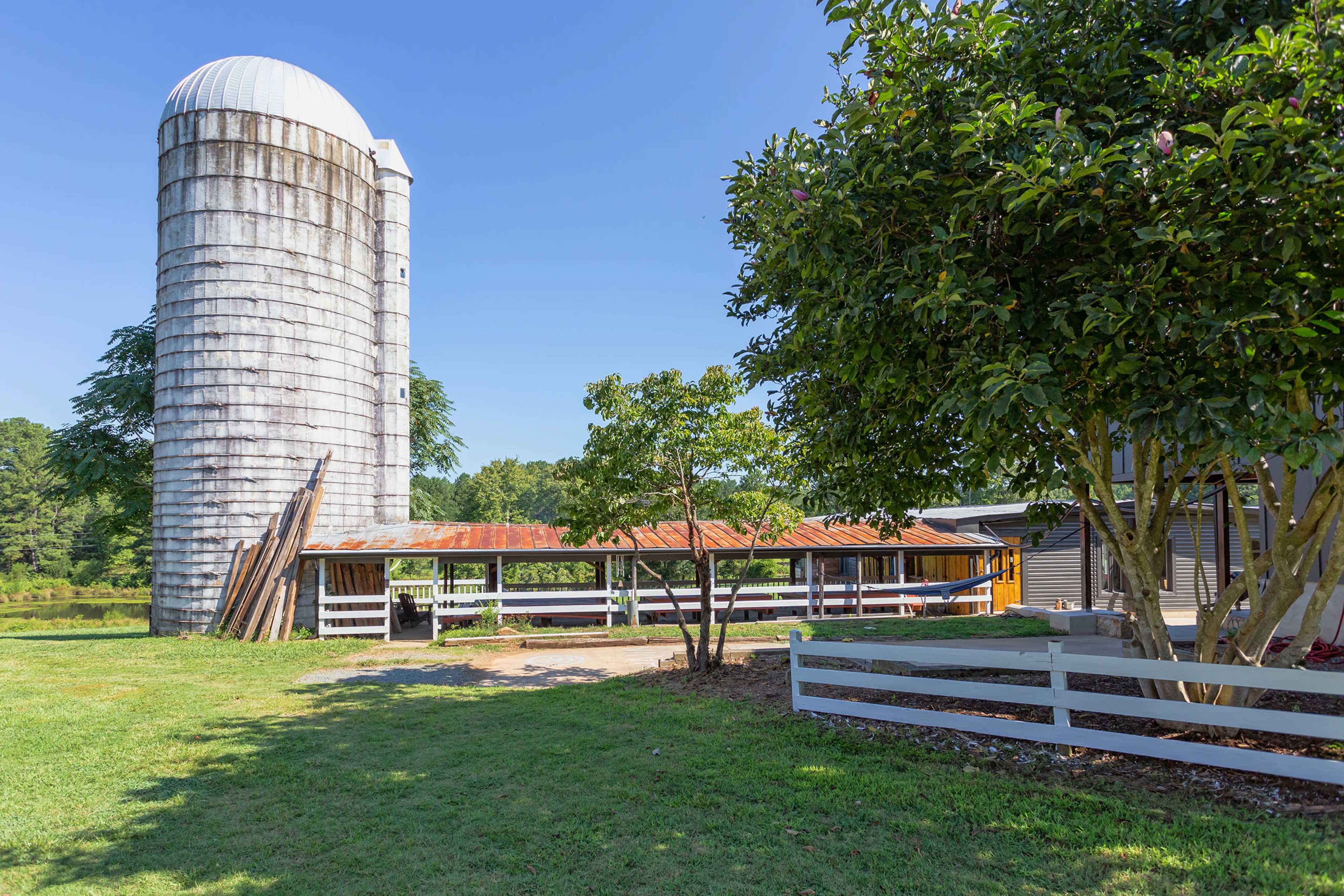 A weathered silo next to an old barn with a rusted roof, surrounded by green grass and a white fence.