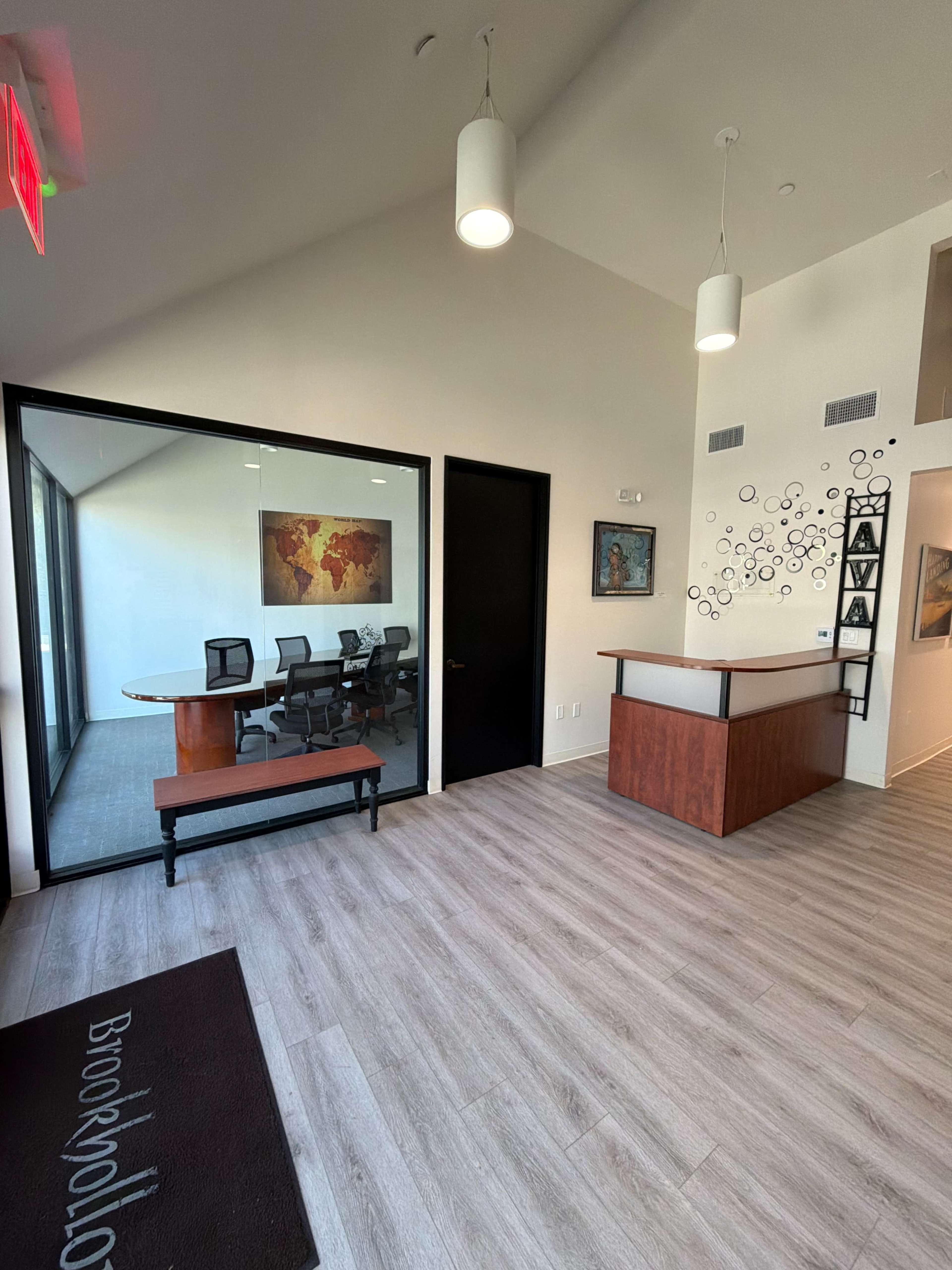 The image shows a modern office reception area with a wooden front desk, a meeting room visible through glass walls, and artwork displayed on the walls.