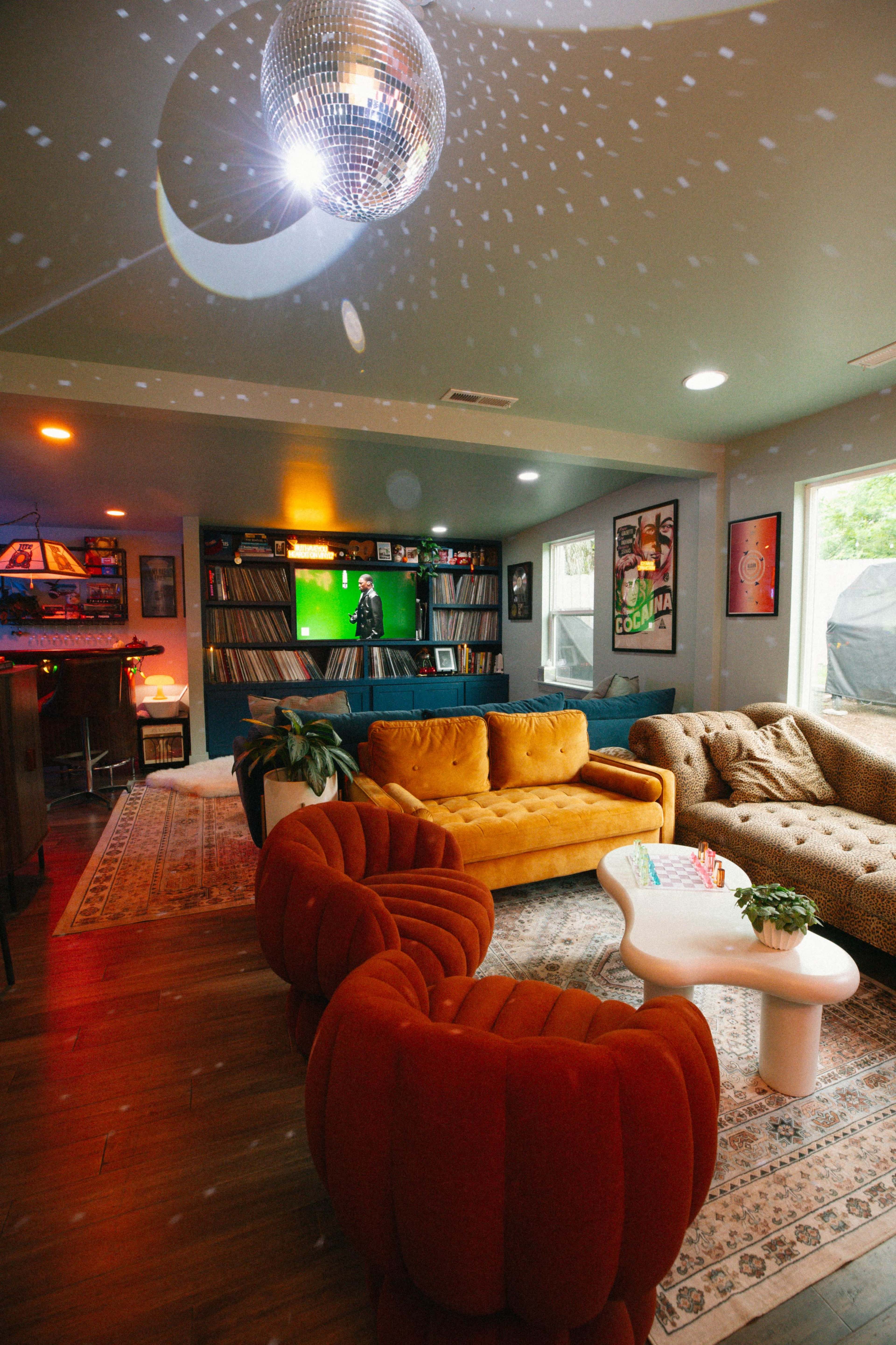 The image shows a colorful living room featuring various seating options, a disco ball, and a wall of vinyl records, with a screen displaying a performance in the background.