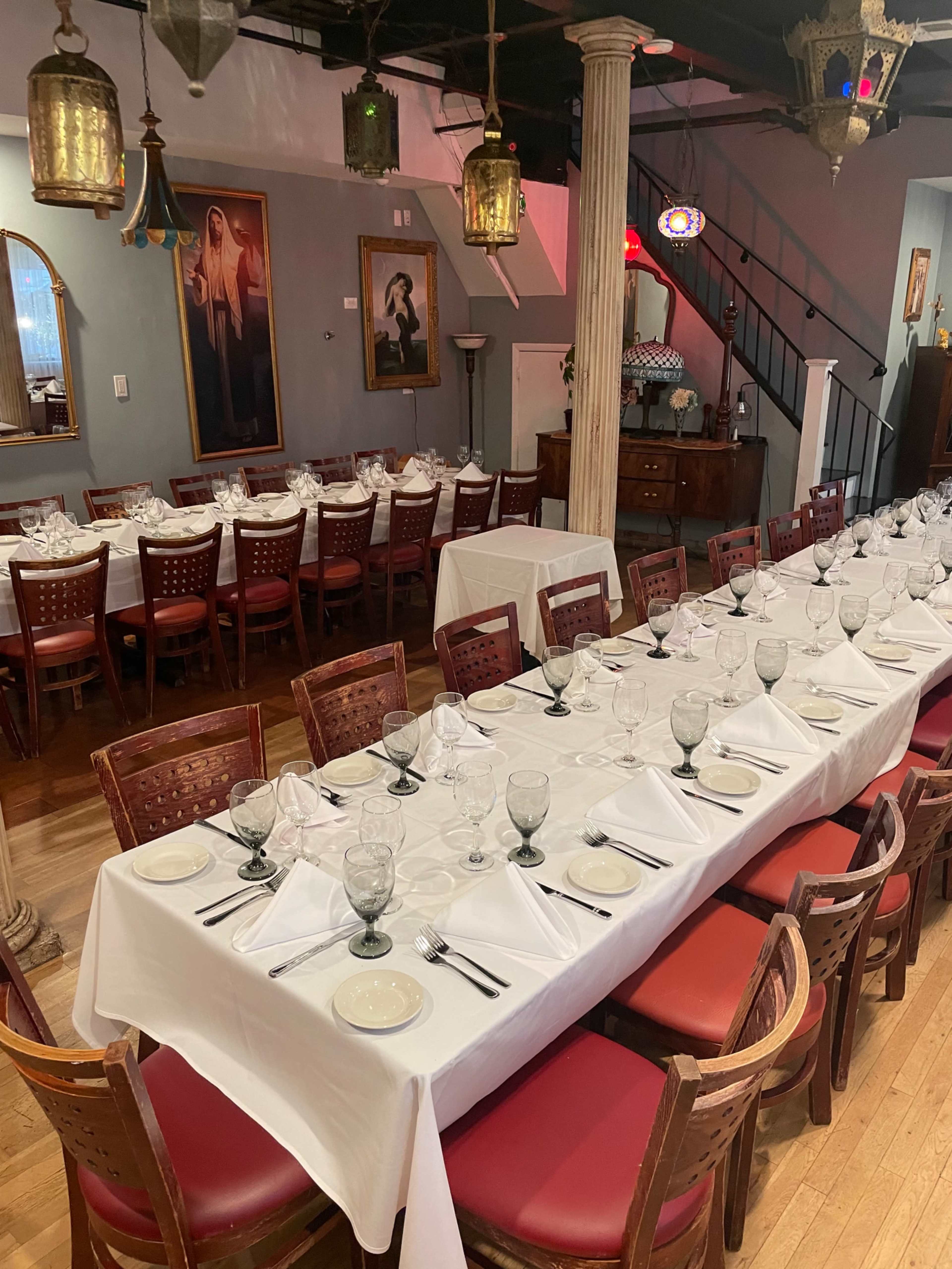 A long dining table set with plates, glasses, and utensils is arranged in an elegantly decorated restaurant featuring paintings and lanterns.