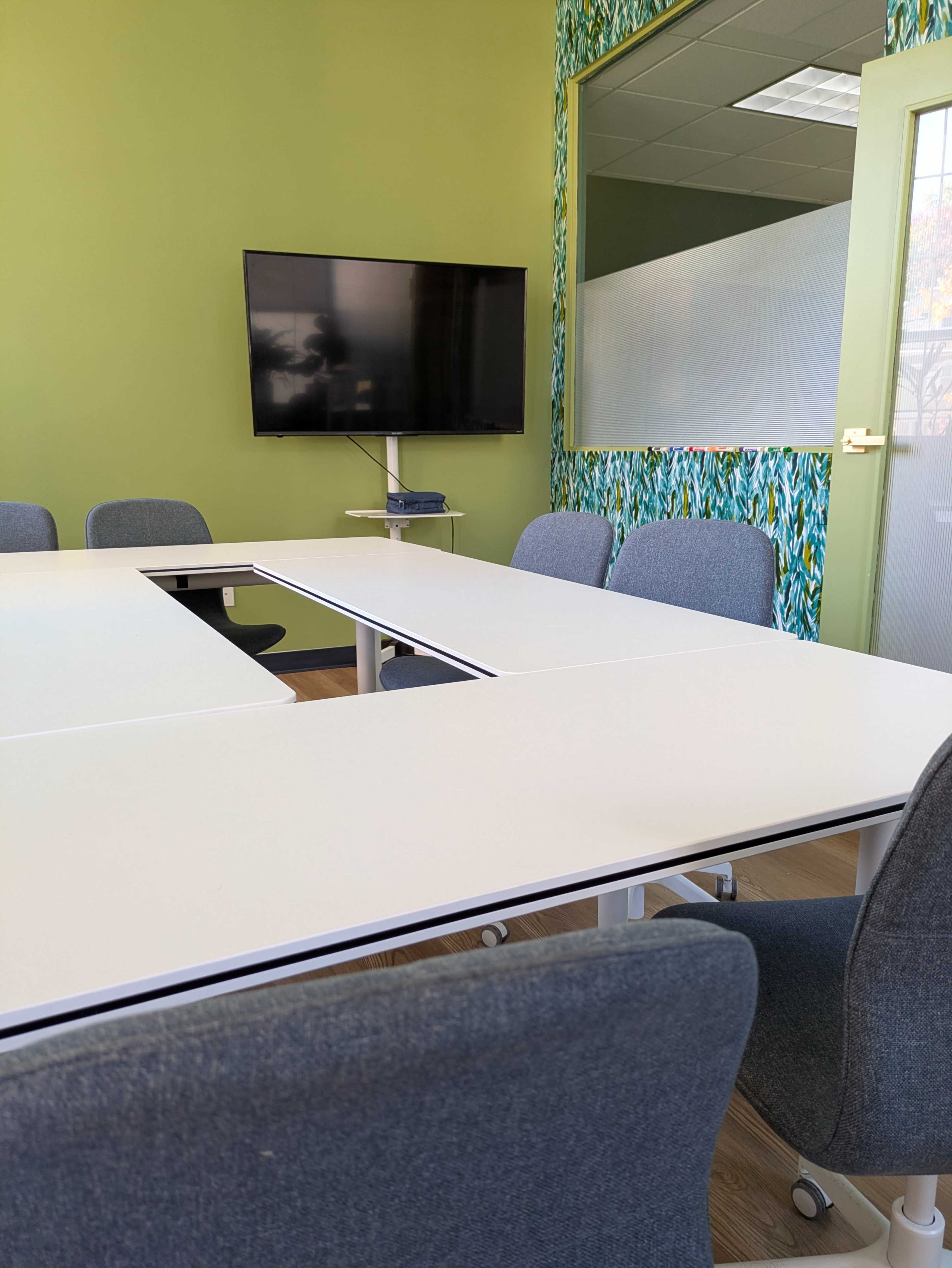 The image shows a meeting room with a rectangular table surrounded by chairs, a television mounted on the wall, and green accent walls with a patterned border.