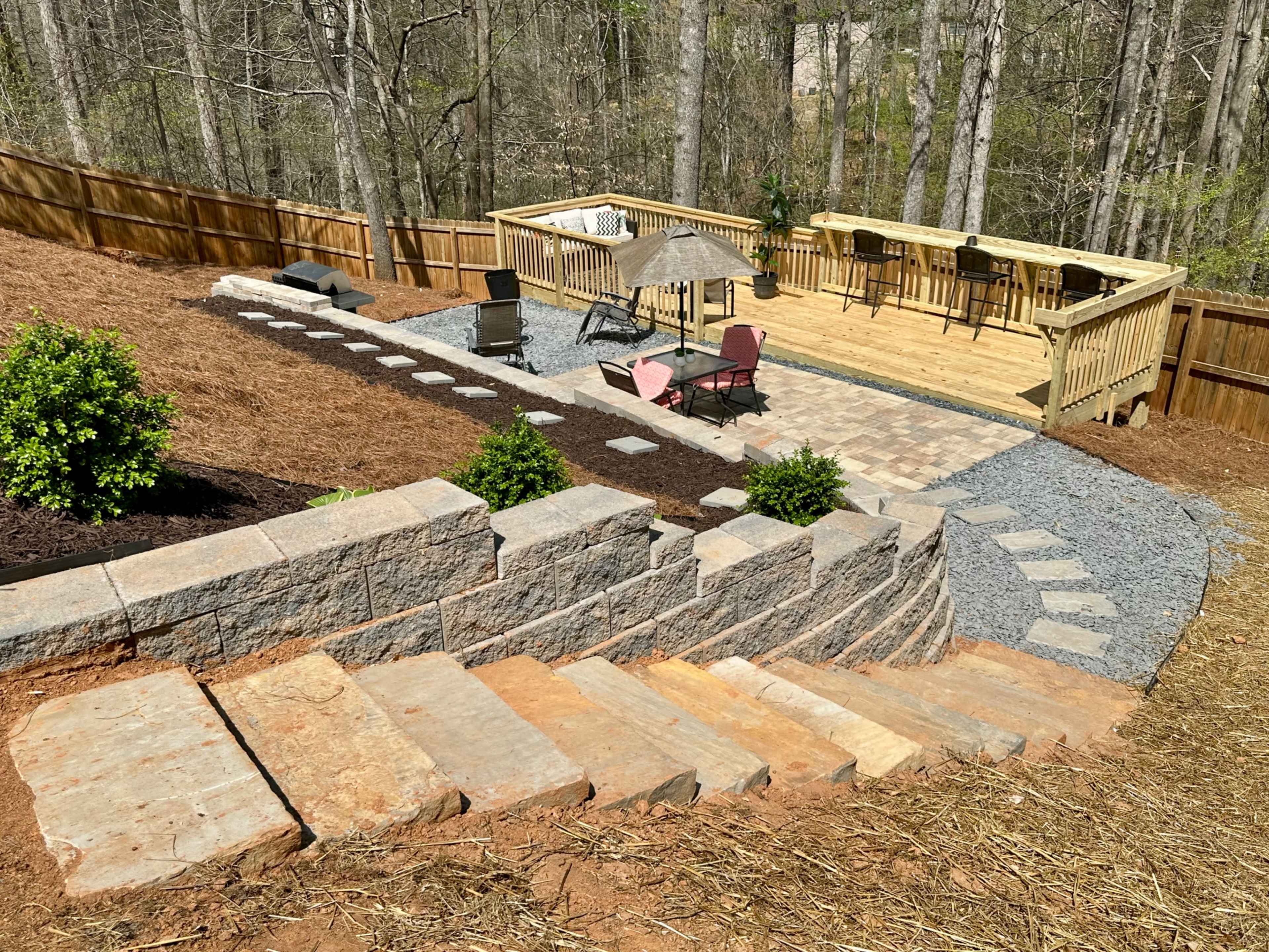 The image shows a landscaped backyard with stone steps leading down to a gravel pathway, a patio area with chairs, and a wooden deck nestled among trees.