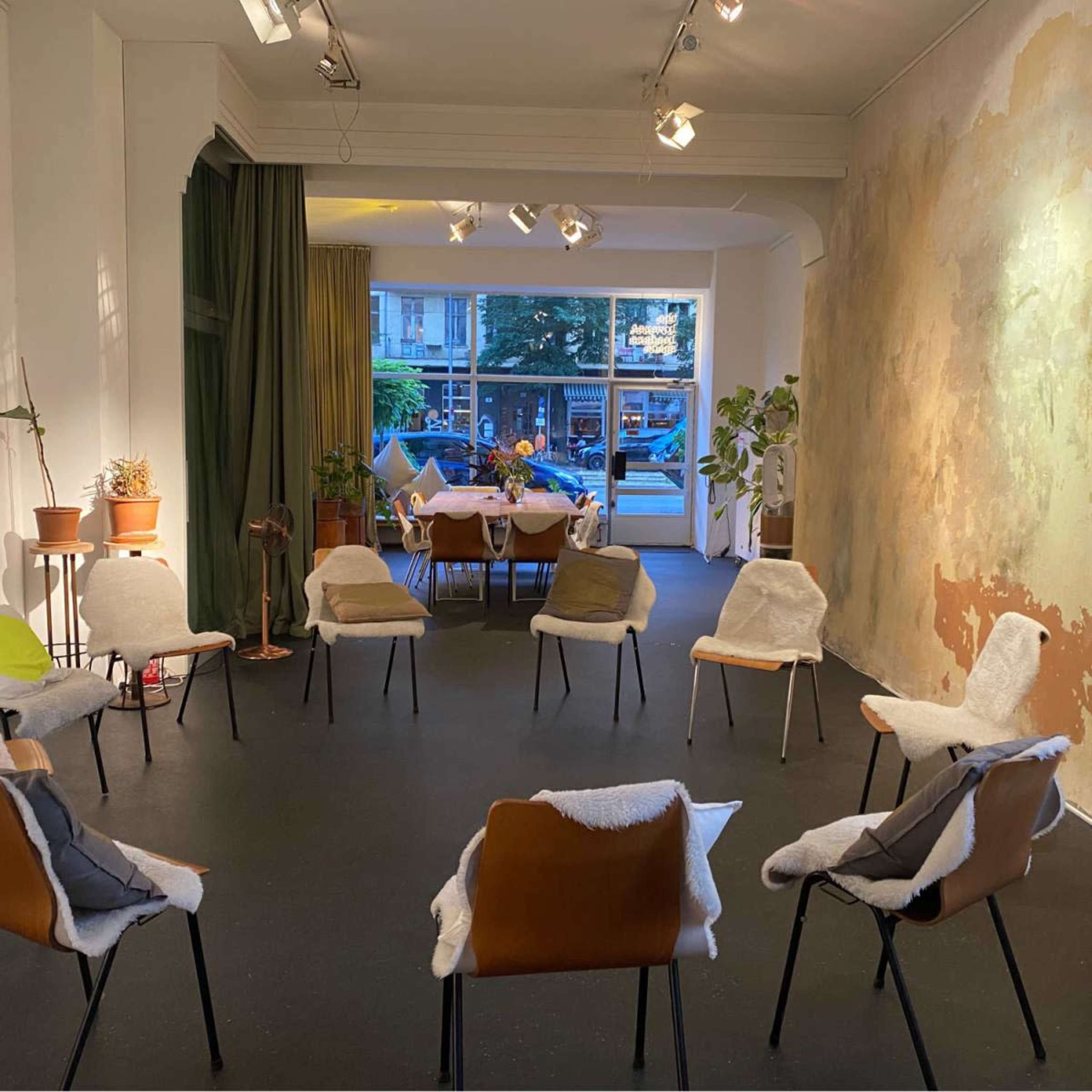 The image shows a room set up with a circular arrangement of chairs, a dining table in the background, and large windows revealing an outdoor view.