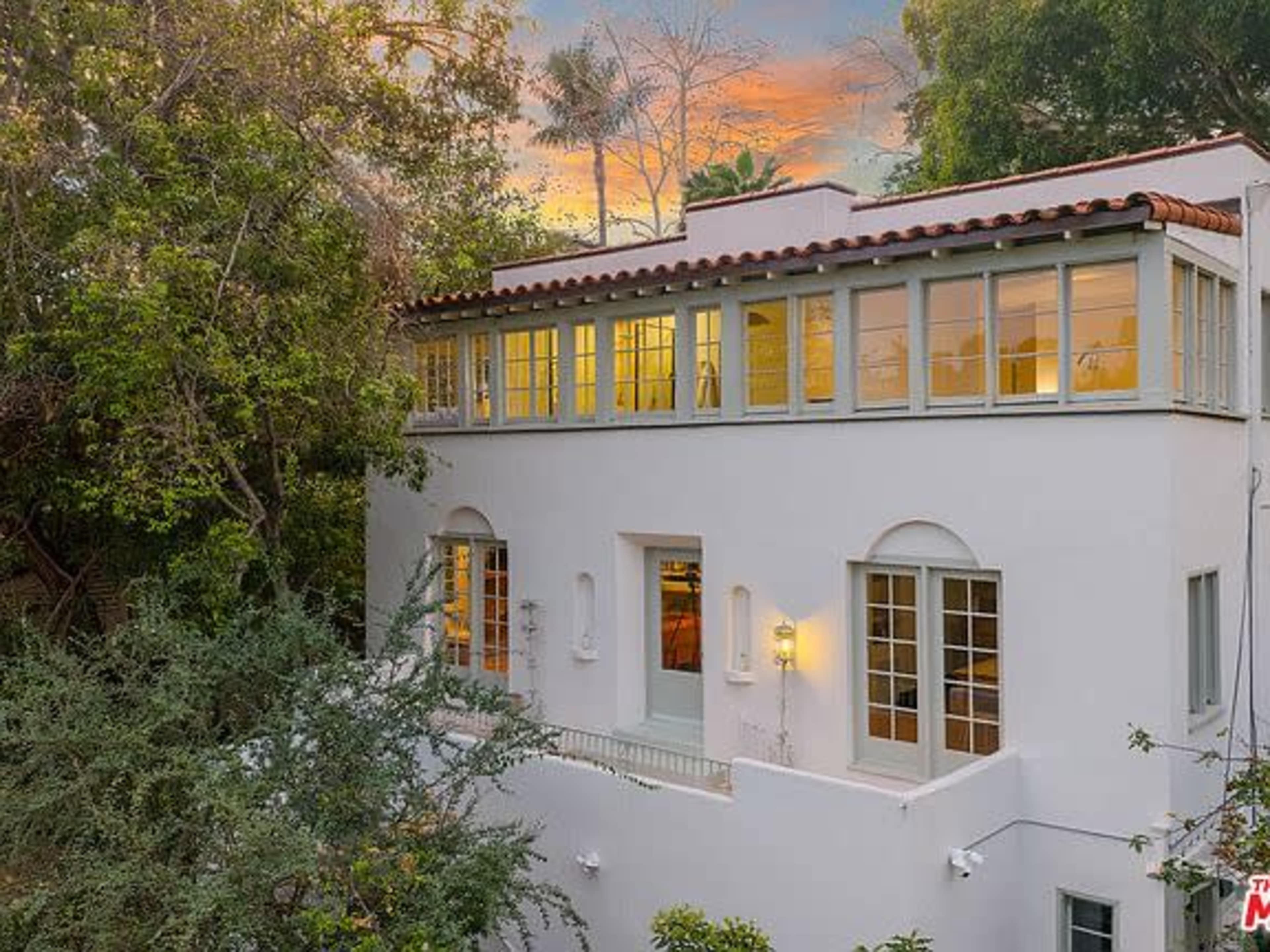 The image shows a white house with multiple windows and a balcony, surrounded by greenery and a colorful sunset in the background.